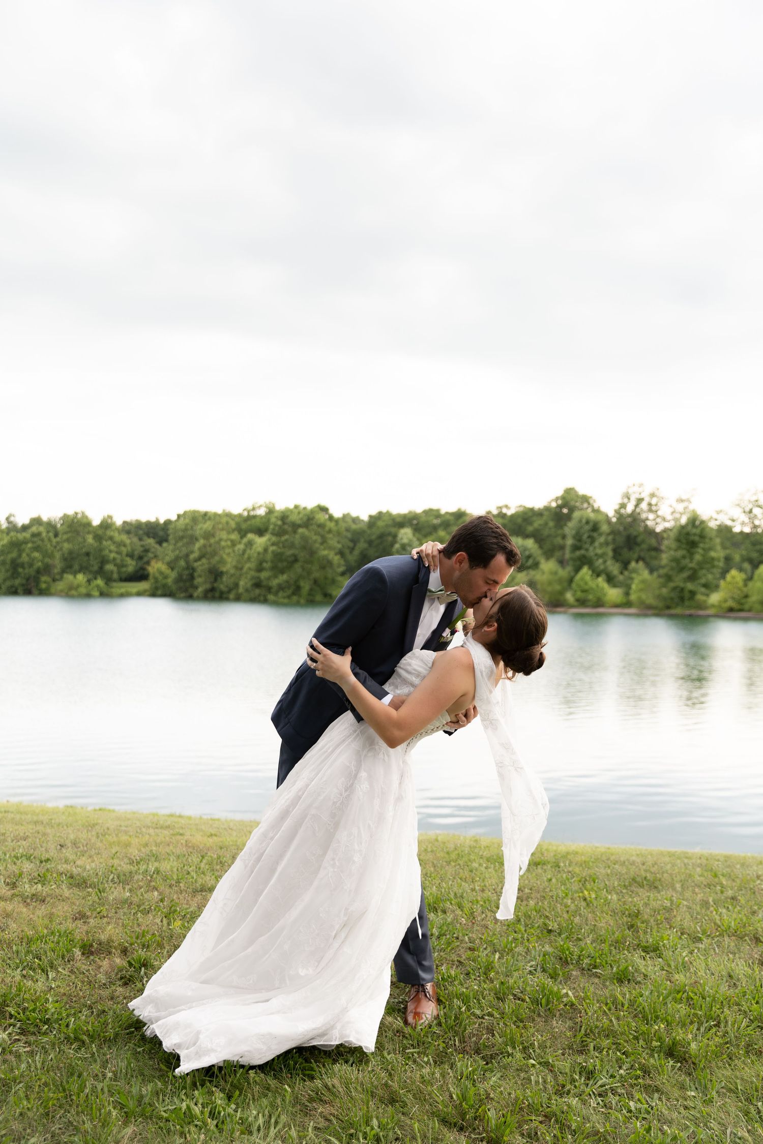 Bride and groom share a romantic dip kiss by a serene lake, surrounded by lush green trees on their wedding day.