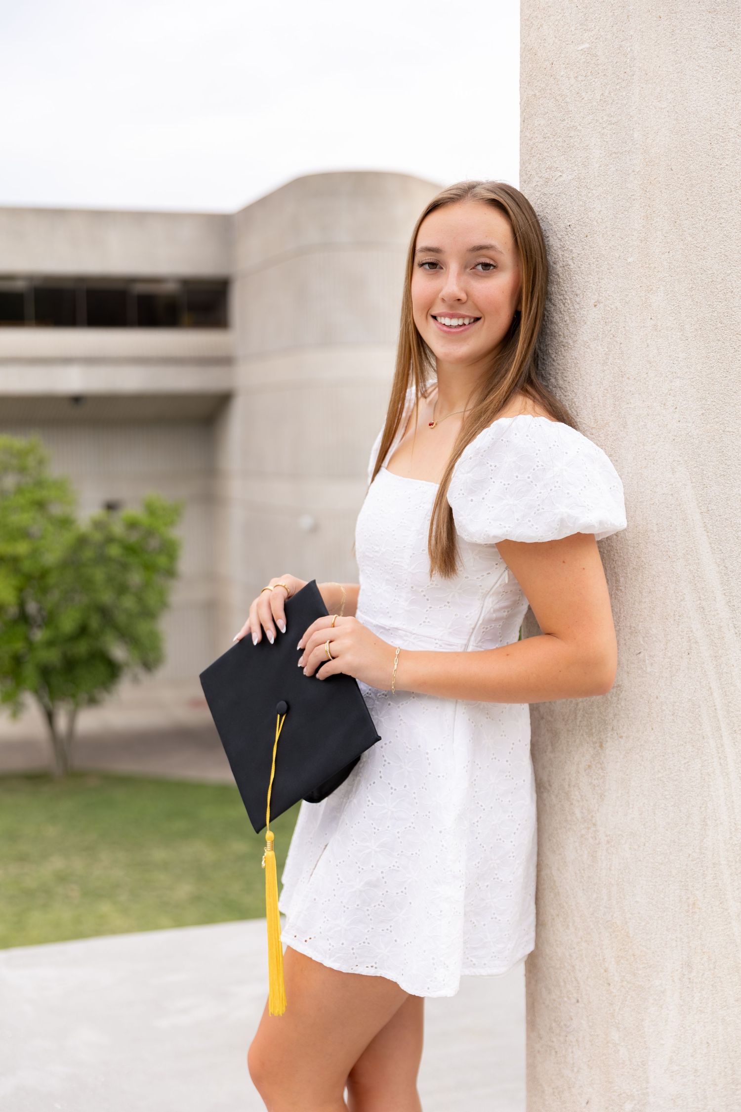 A smiling young woman in a white dress holds her graduation cap against a stone wall.