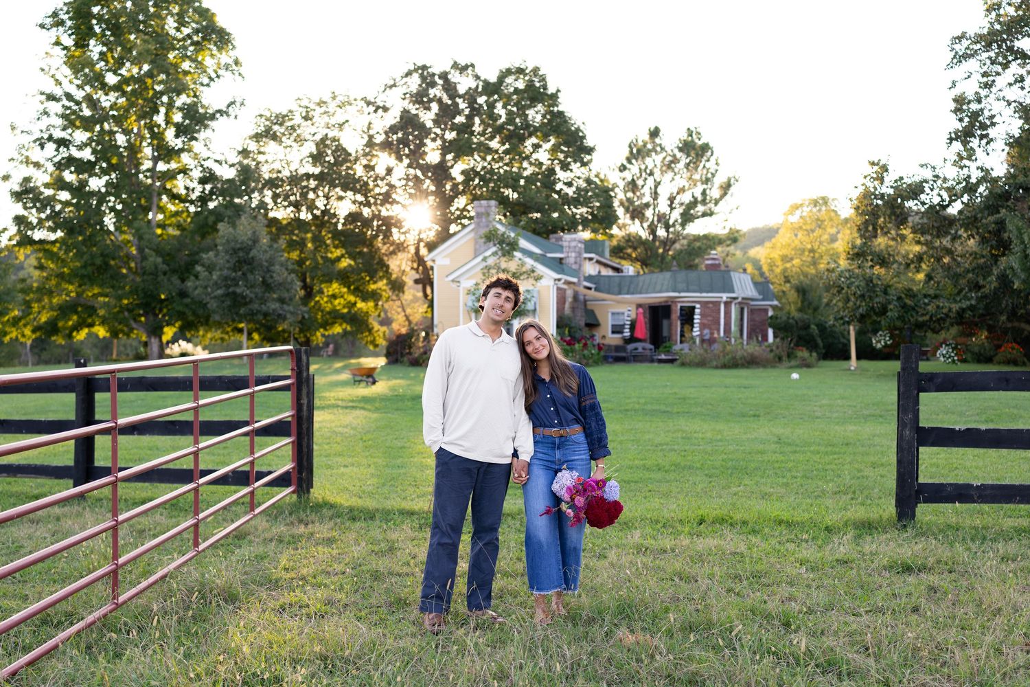 Couple posing together in front of a farmhouse at golden hour, surrounded by green fields and wooden fencing.