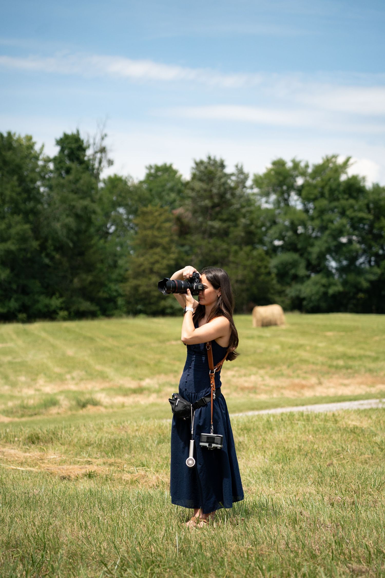 Woman in a navy dress photographing in a green field with trees and a hay bale under blue sky.