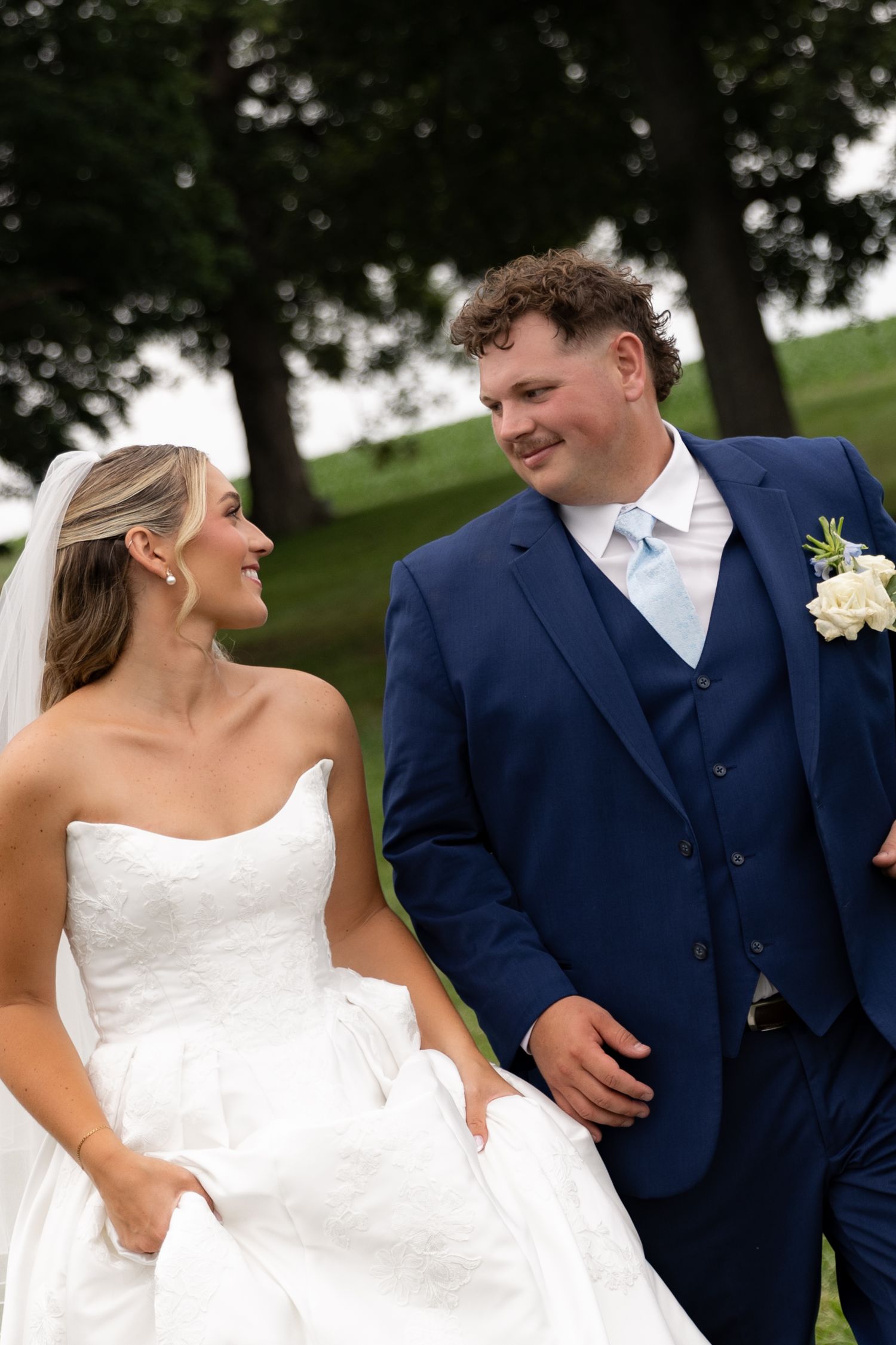 Bride in white strapless gown and groom in navy blue suit smile at each other outdoors on their wedding day.