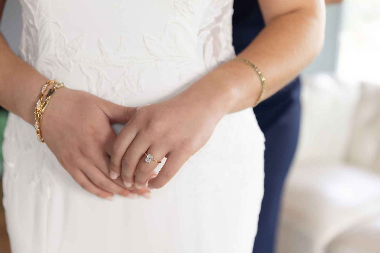 Bride in white dress with diamond engagement ring and gold bracelet, hands clasped at waist.