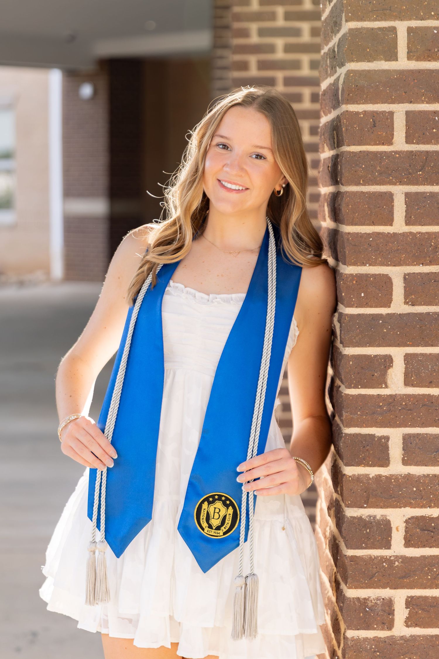 Smiling young woman in white dress wearing blue graduation stole with gold emblem, leaning against brick wall outdoors.