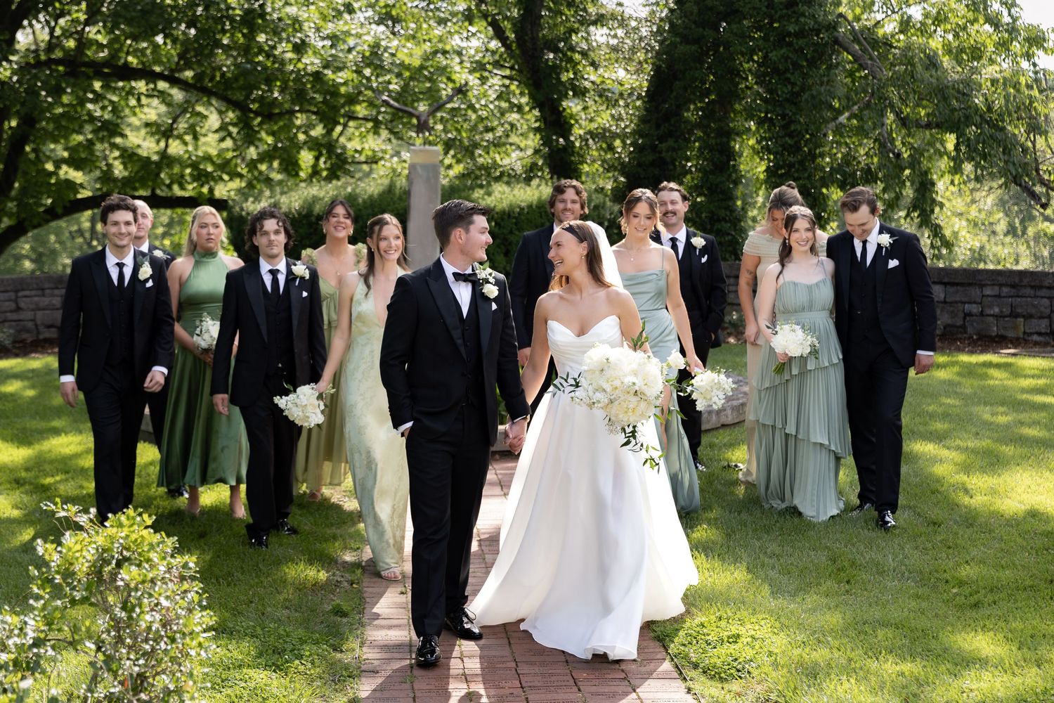 Wedding party walks on brick path, bride in white gown holding bouquet, groom in tuxedo, attendants in sage green and black.