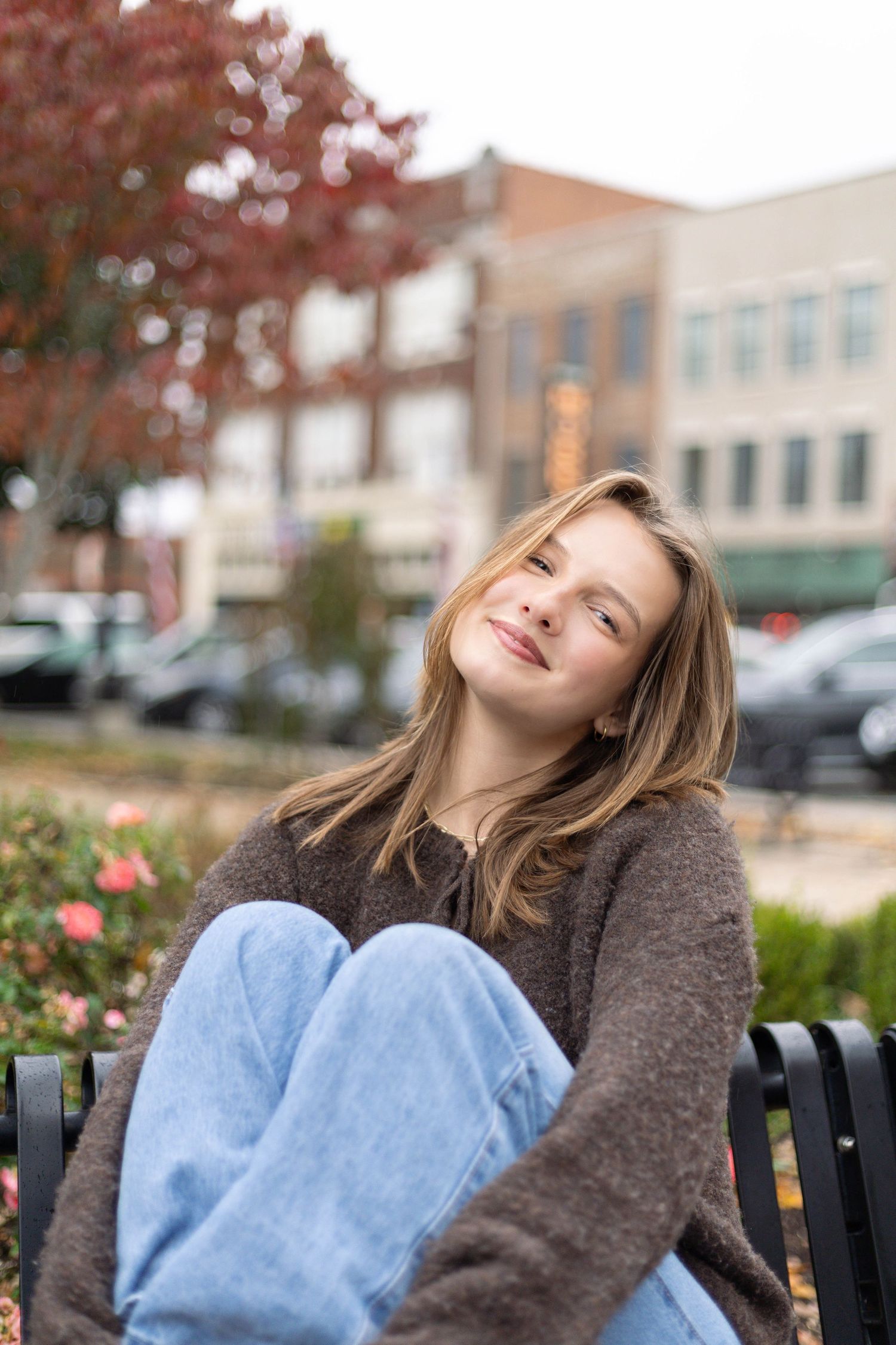 Teenage girl in brown sweater and jeans sits on bench smiling in urban fall setting.