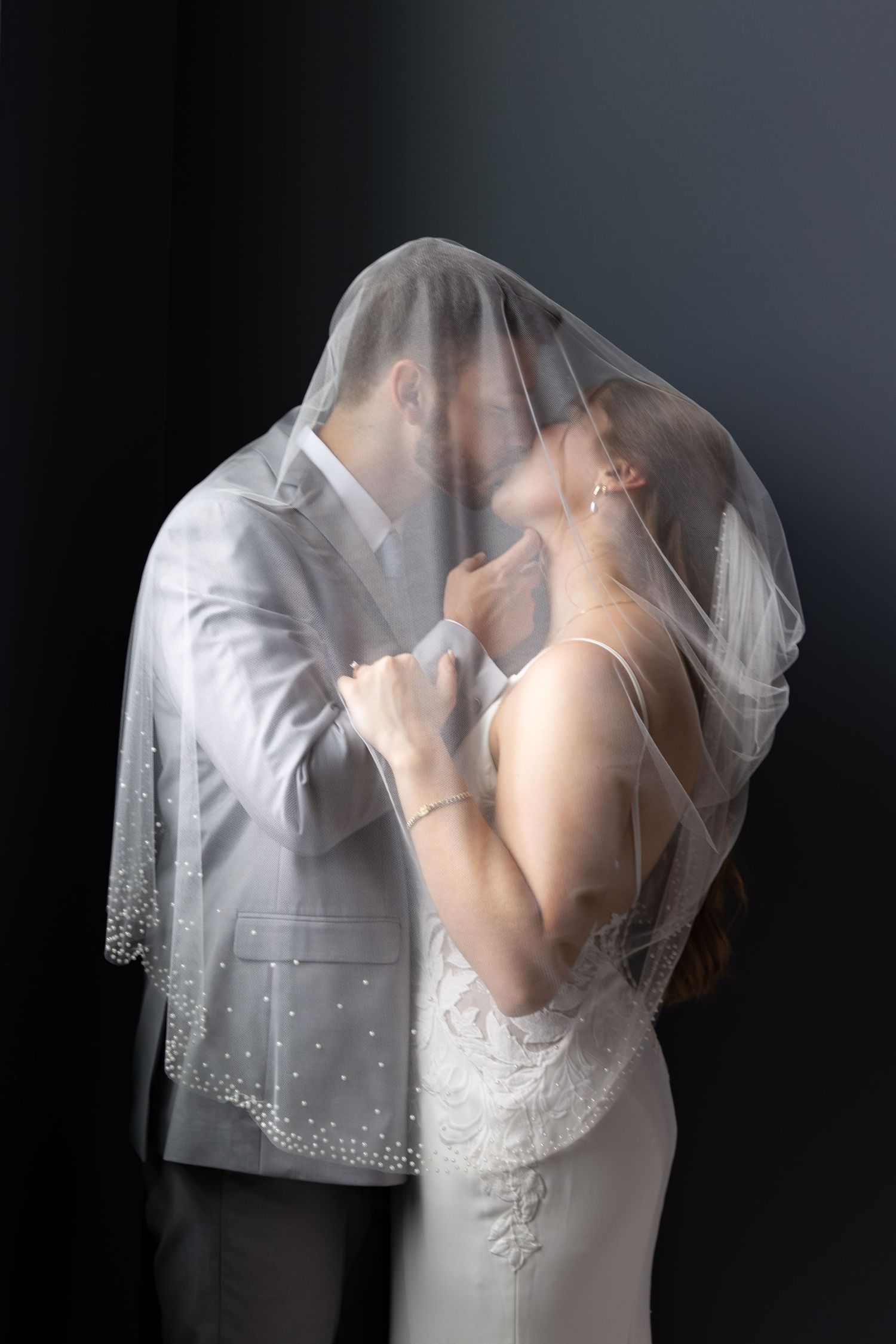 A bride and groom share a romantic kiss under a pearl-embellished wedding veil against a dark background.