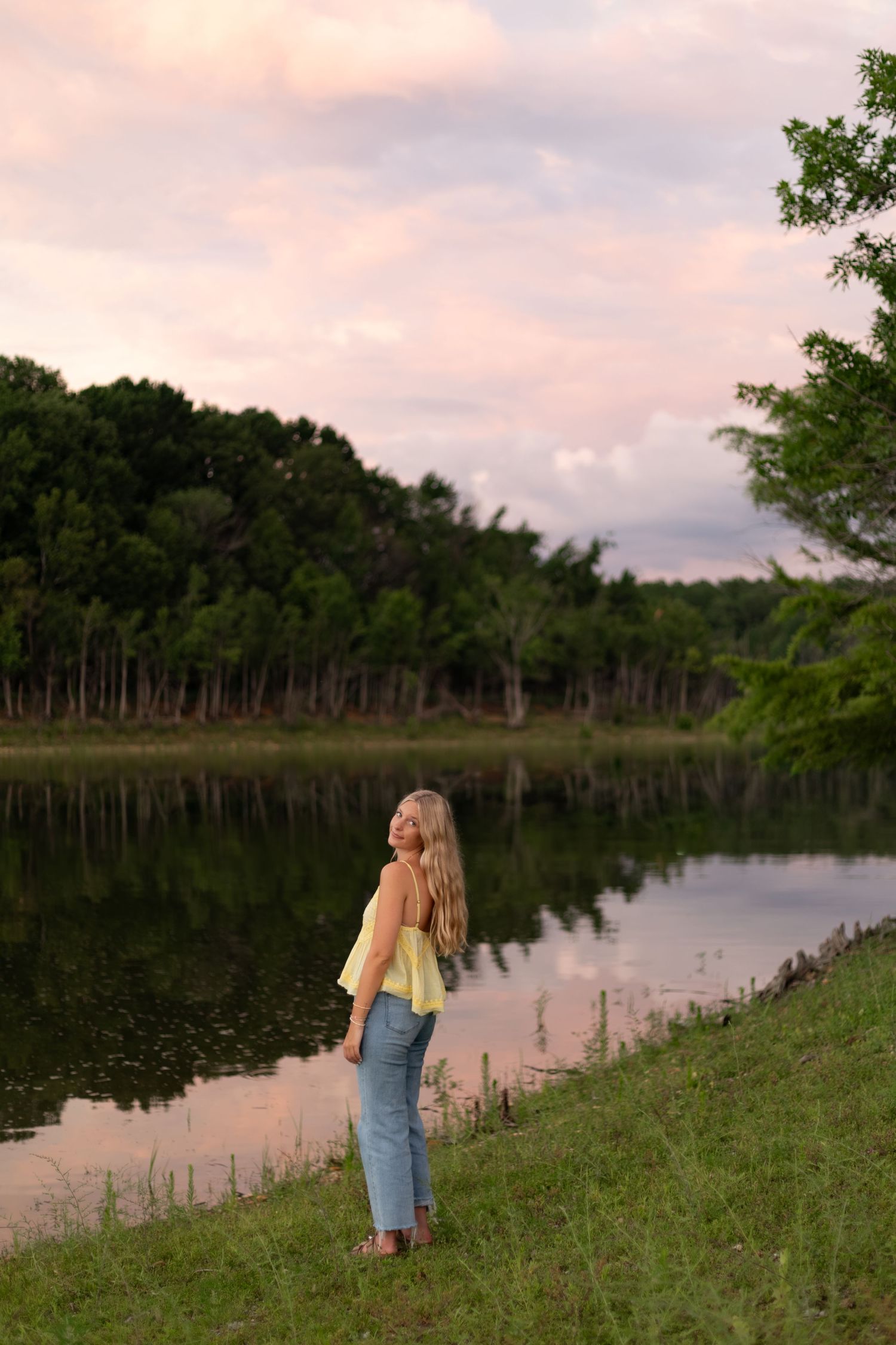 Blonde woman in yellow top and jeans standing by a serene lake at sunset with pink clouds and tree reflections.
