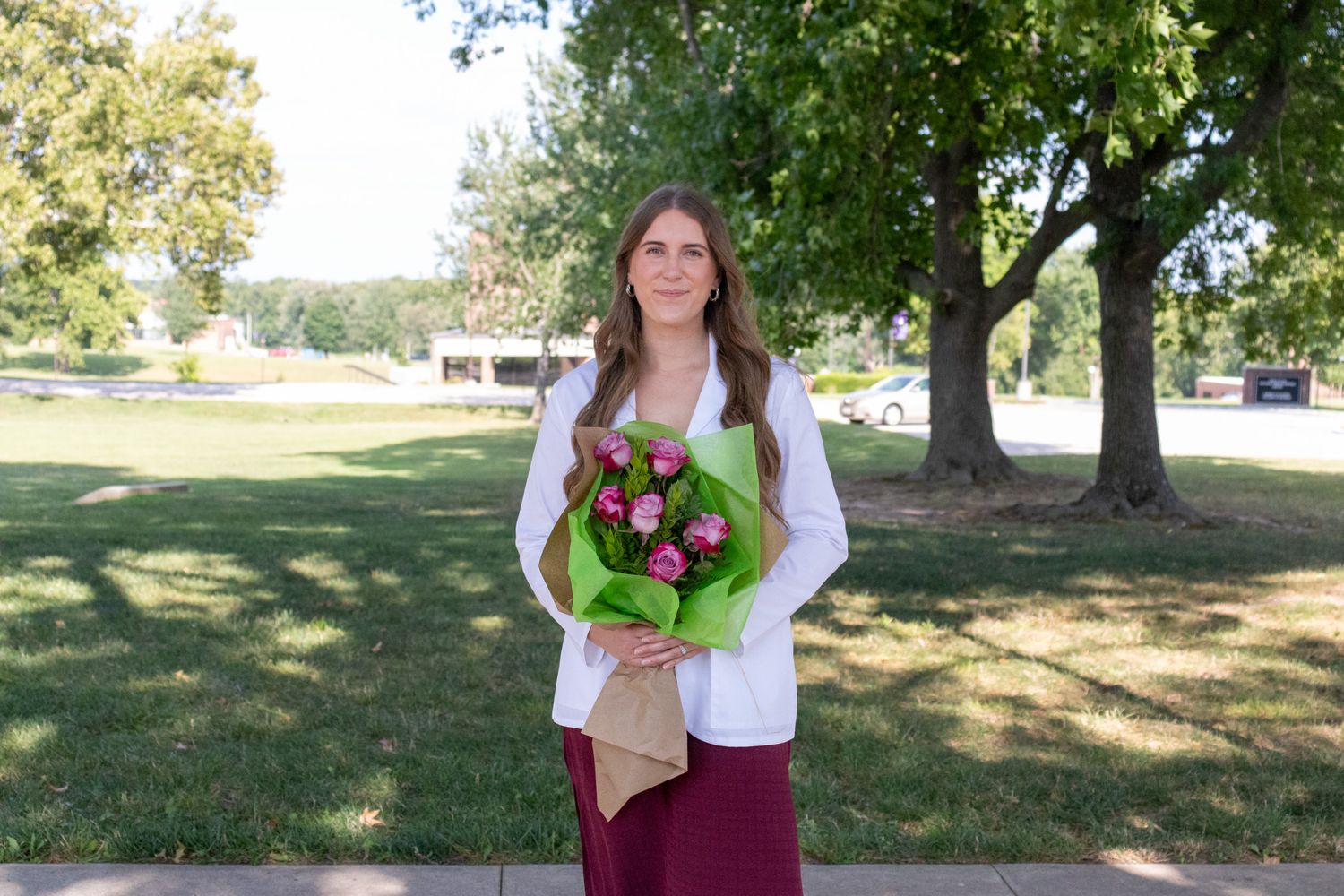 Woman in white coat holding pink flower bouquet standing on a sunny campus.