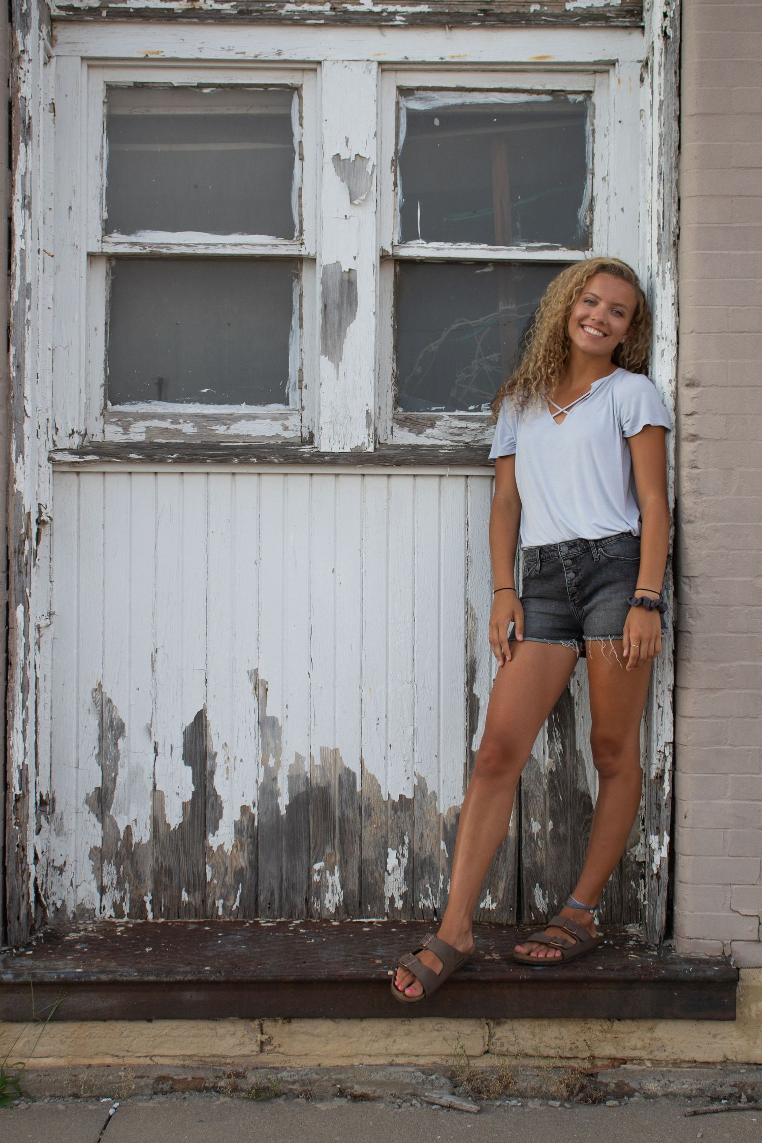 Smiling teenage girl in white tee and denim shorts posing by rustic door.