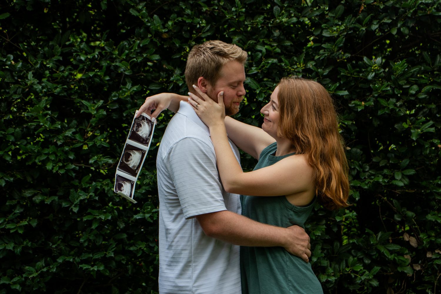 Couple embracing outdoors holding ultrasound photos in front of green hedge.