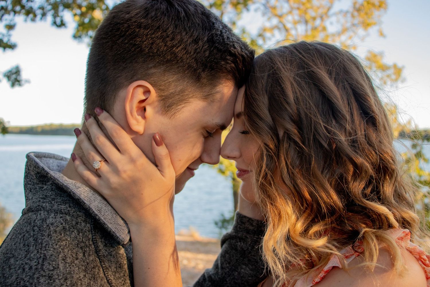 Romantic couple touching foreheads near a lake surrounded by autumn foliage.