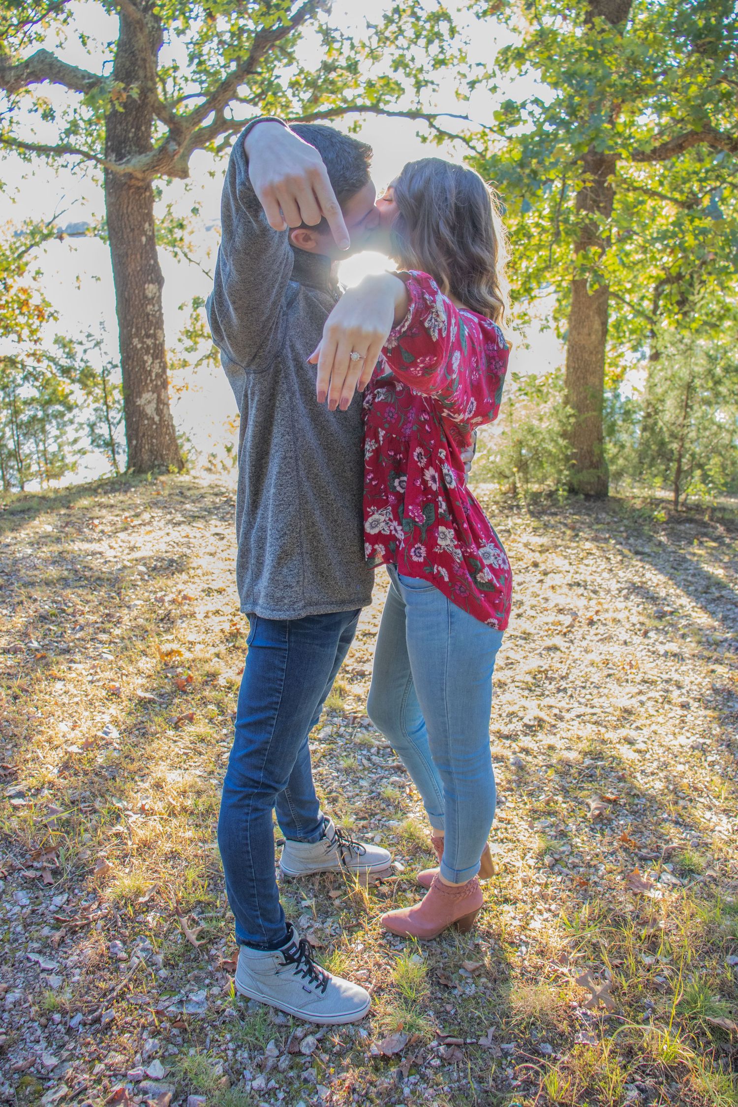 Engaged couple kissing outdoors showing off engagement ring in sunlit forest.