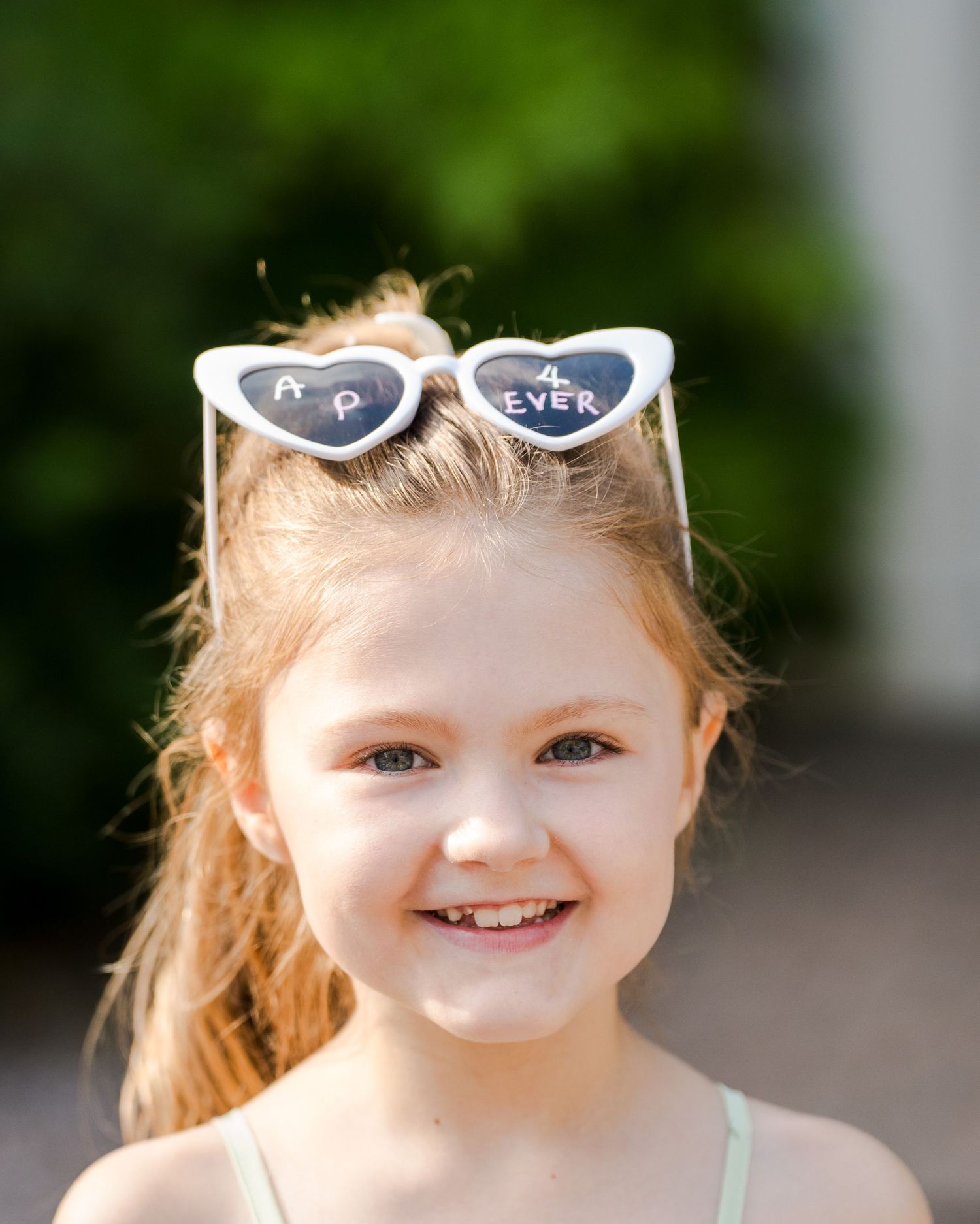 A smiling young girl with heart-shaped cat-eye sunglasses pushed up on her head at a sunny wedding.