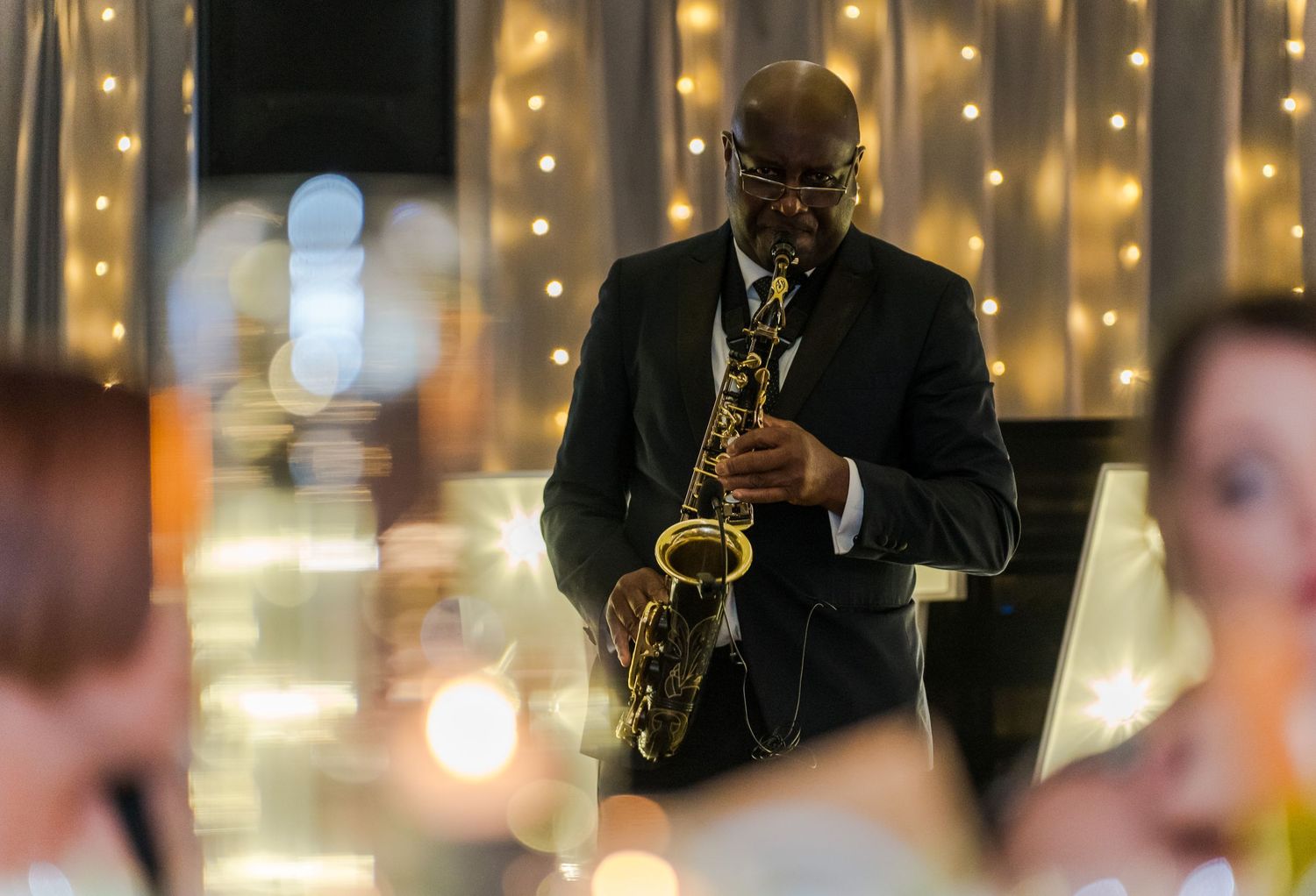 A saxophonist performs in formal attire at an elegant wedding reception with warm fairy light backdrop.