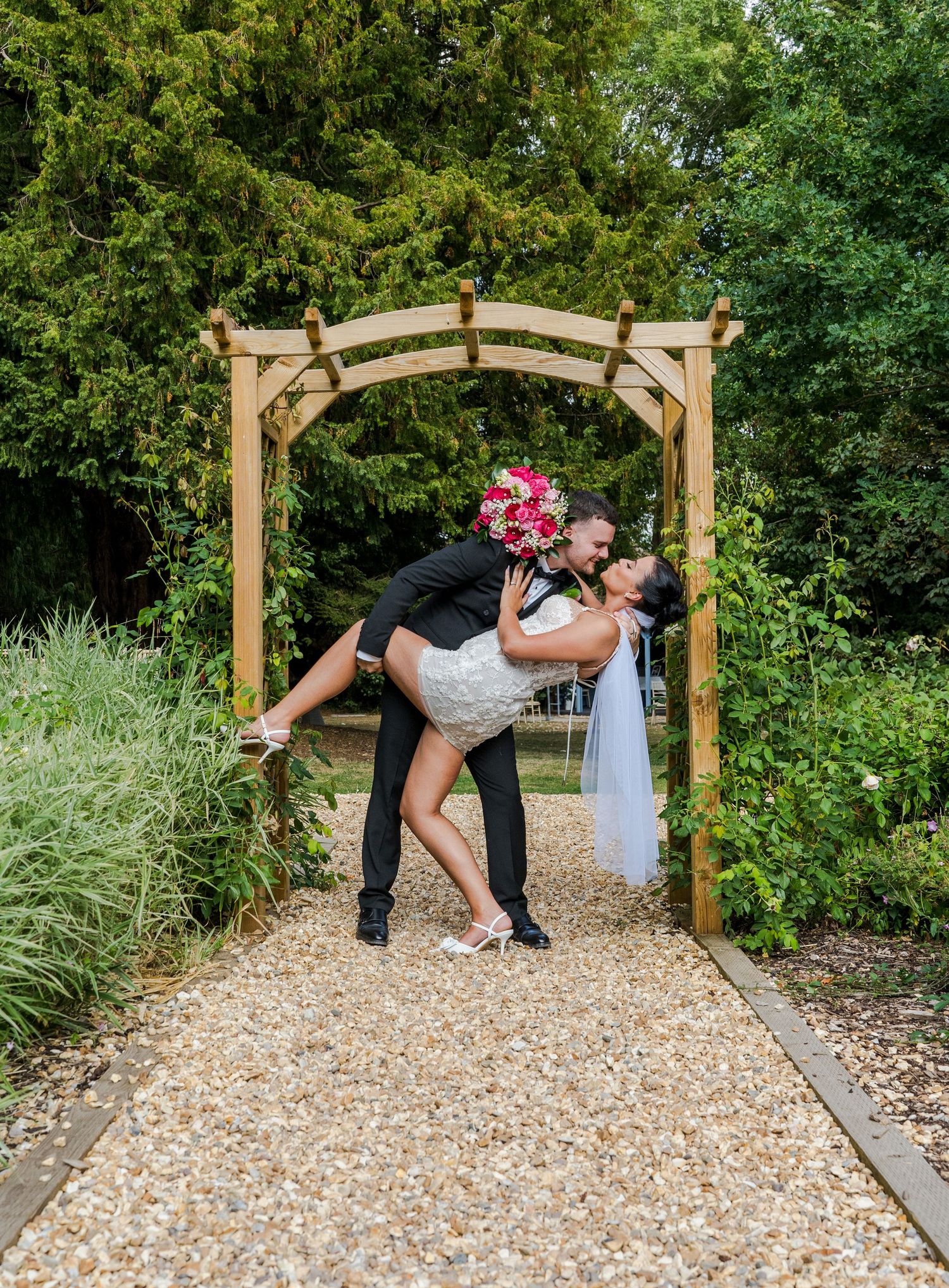 Groom dips bride in elegant white gown under wooden arch on gravel garden path at wedding.