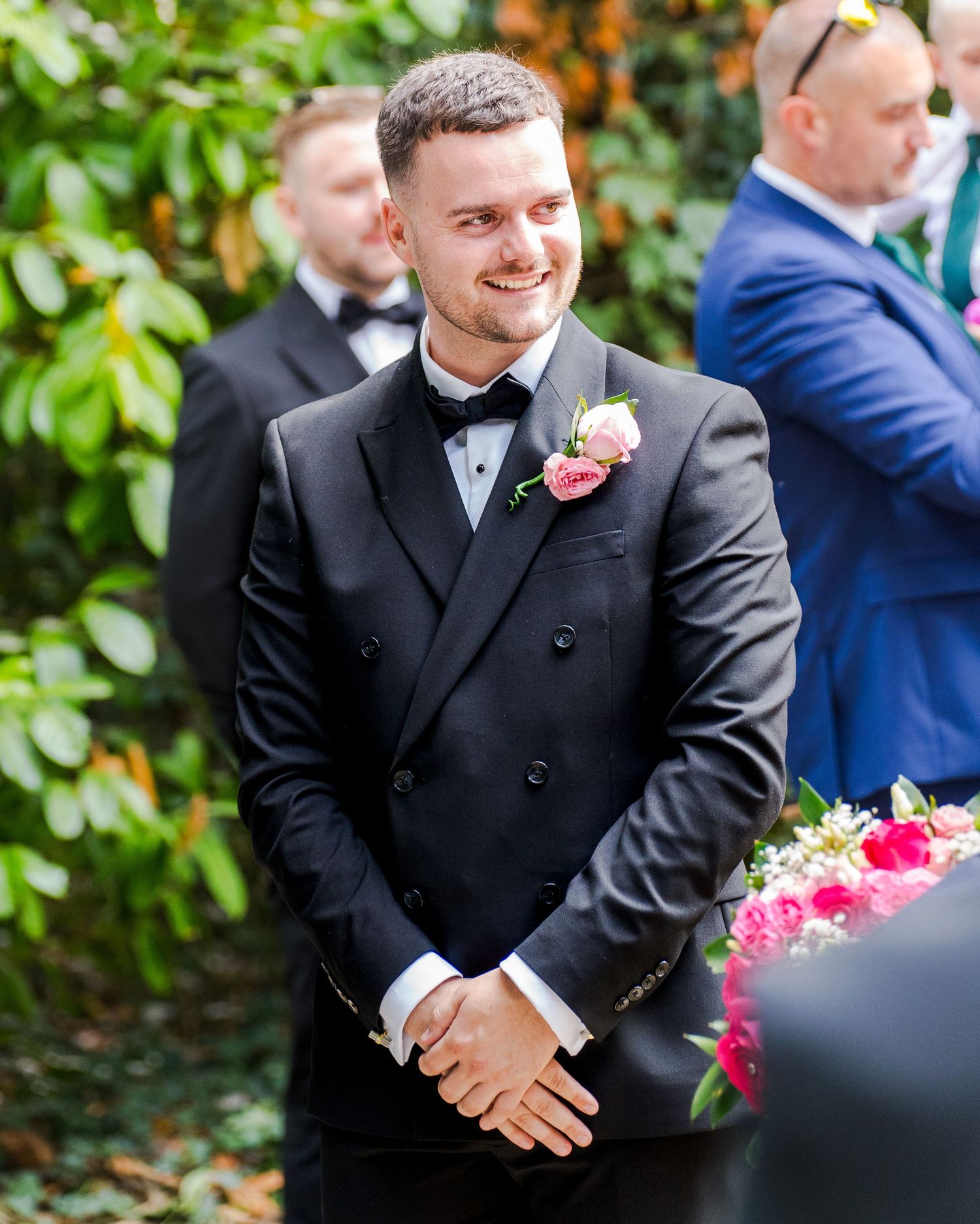 Smiling groom in black tuxedo with pink boutonniere at outdoor wedding ceremony.