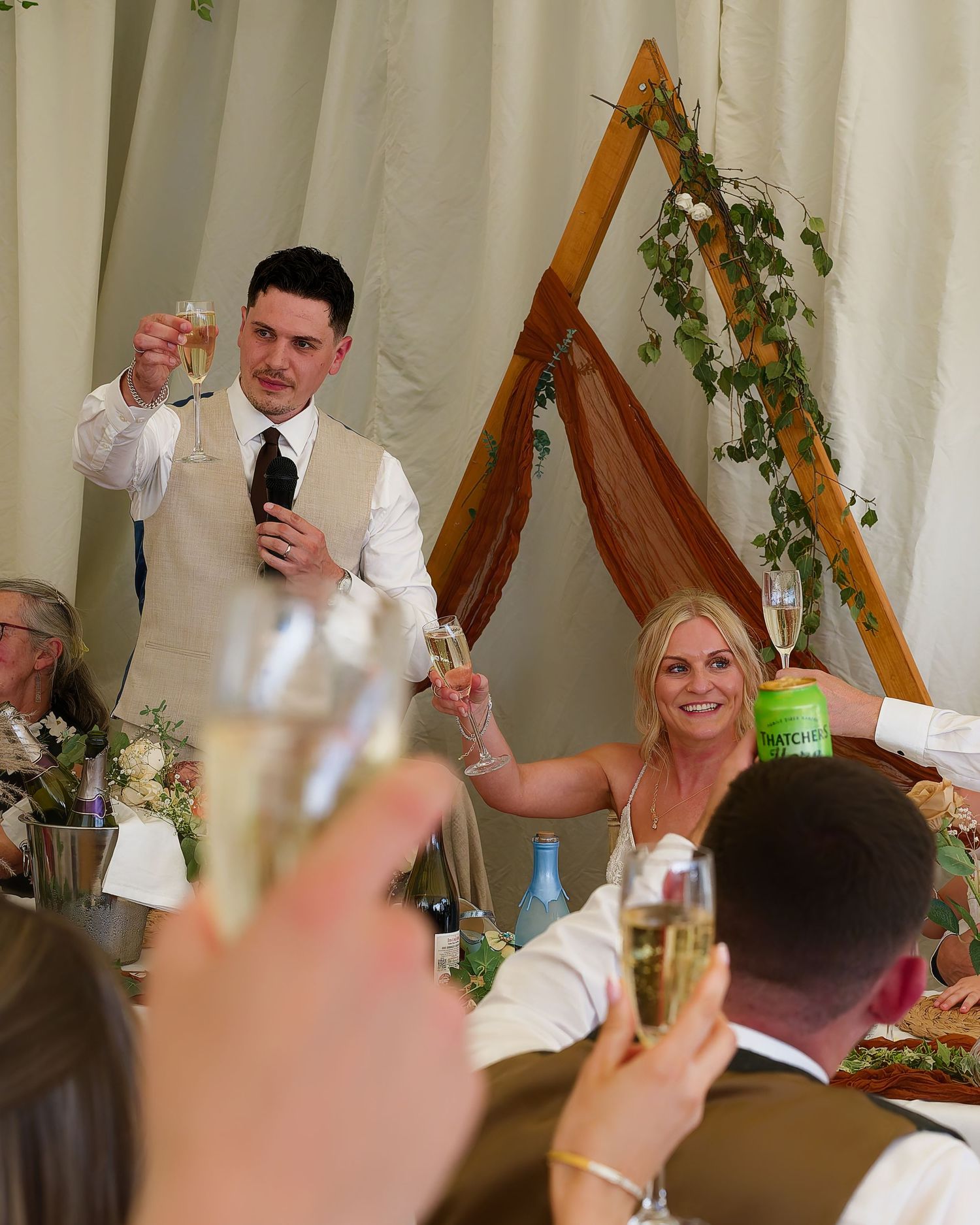 Man in vest raises champagne glass giving wedding toast as blonde woman smiles and lifts her glass at reception.