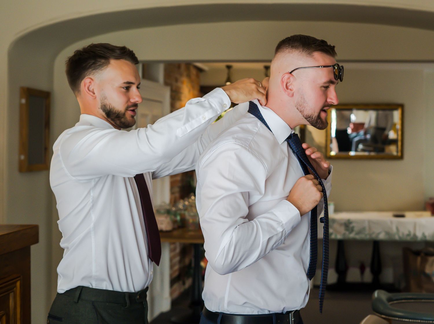 Groomsman helping the groom straighten his tie while getting ready indoors before the wedding.