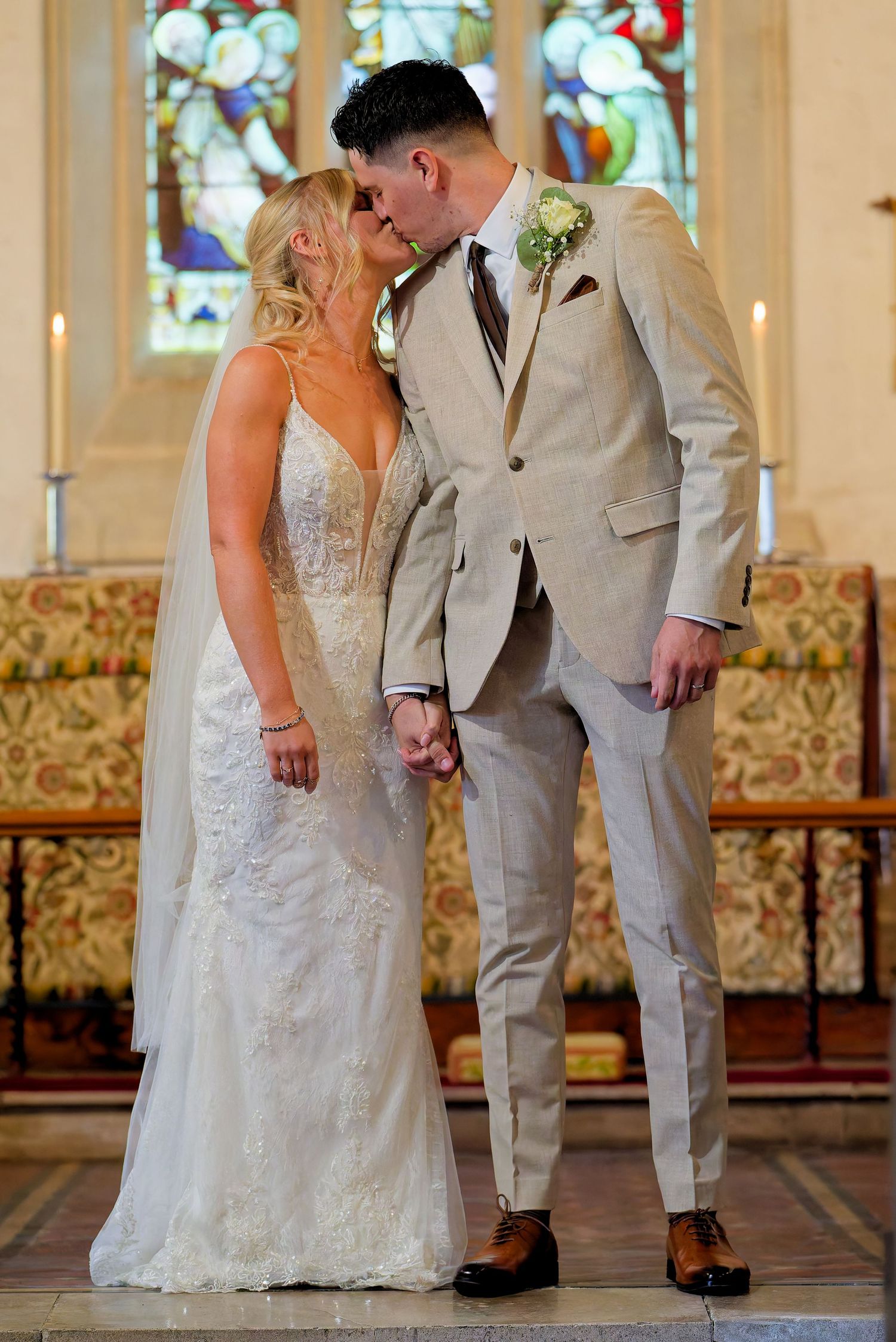 Bride and groom share first kiss at church altar, groom in beige suit, bride in lace wedding gown.