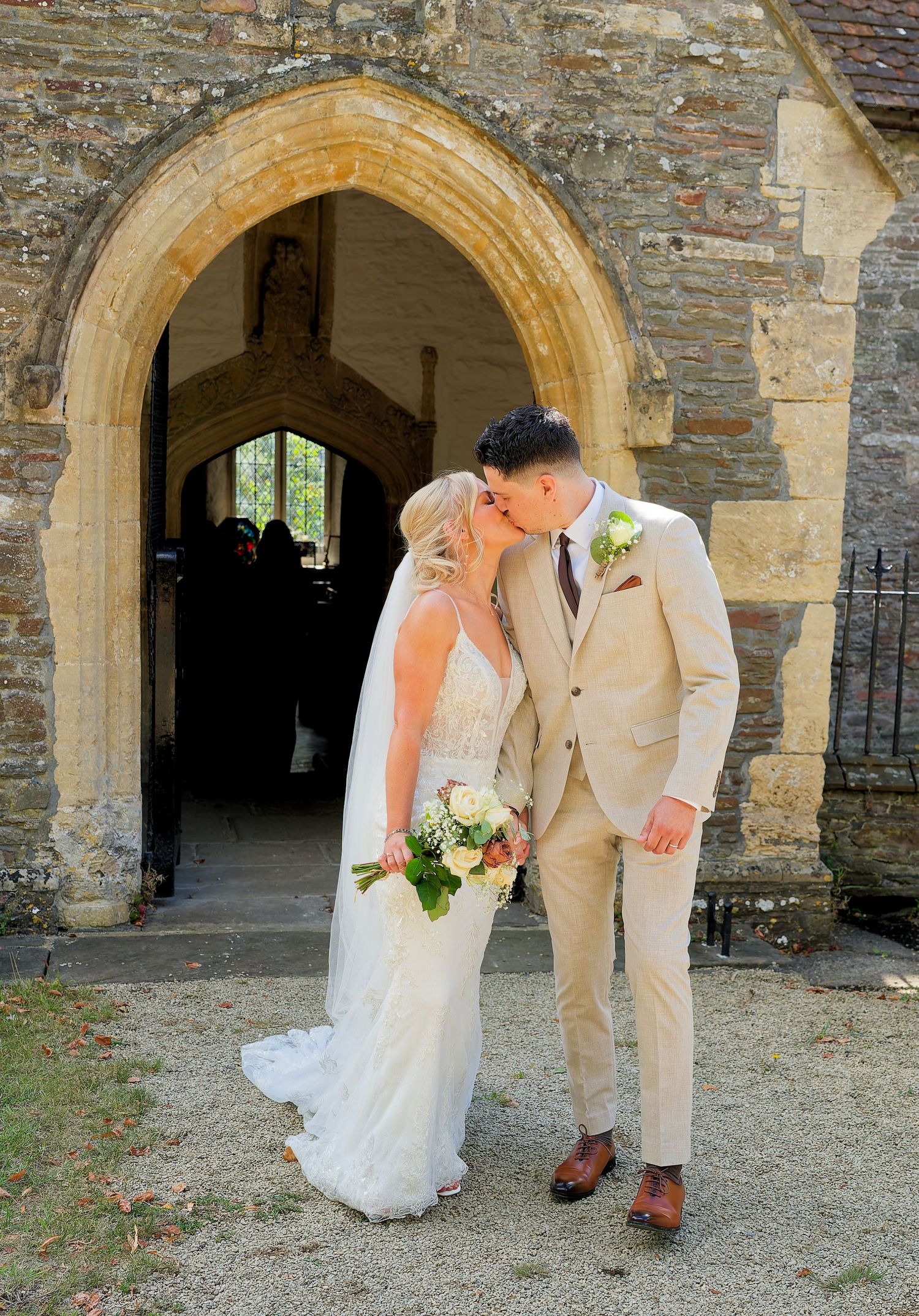 Bride and groom kissing under stone archway of historic church after wedding ceremony.