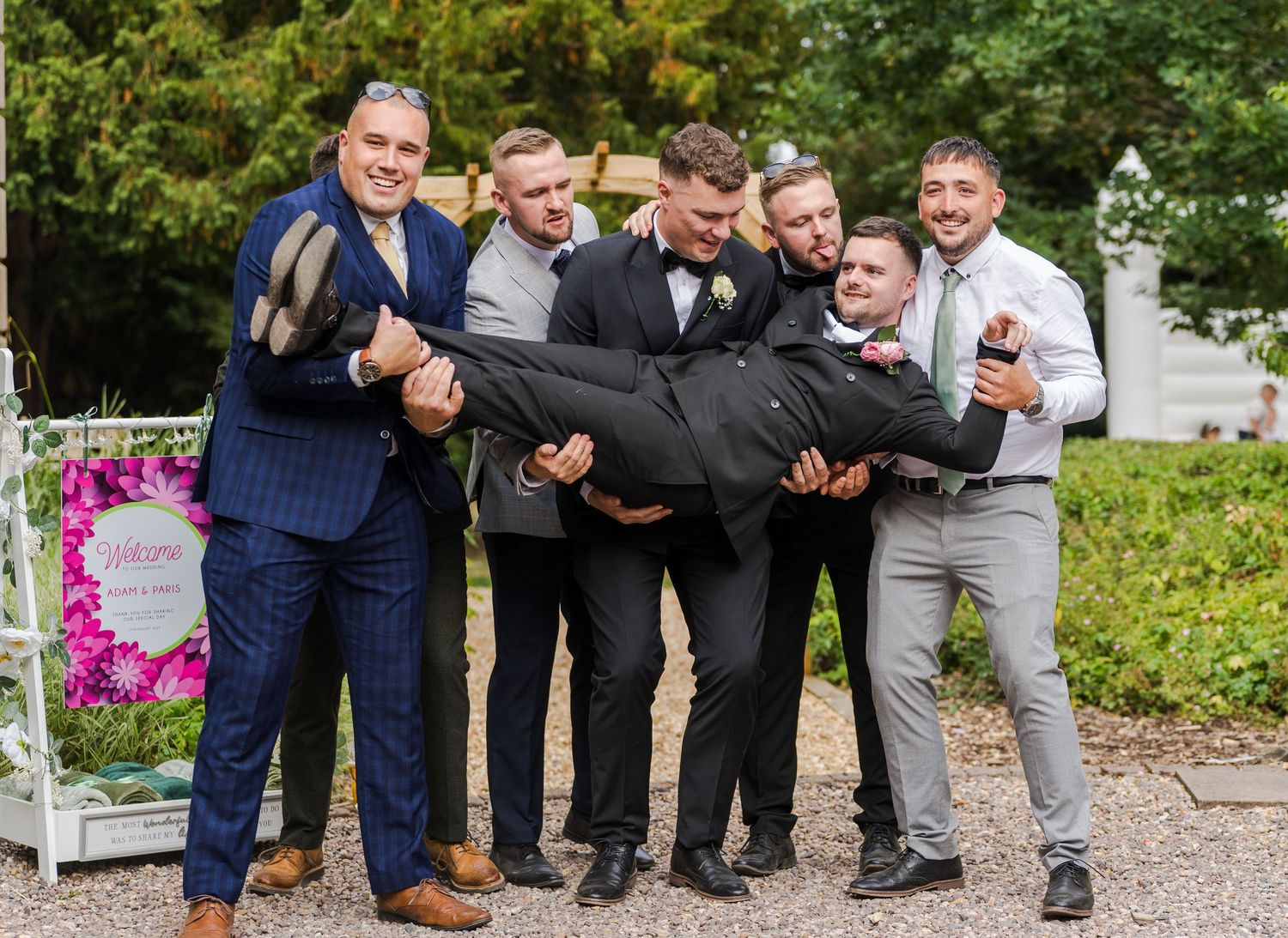 Groomsmen playfully carry groom in black tuxedo outside wedding venue in fun group photo.