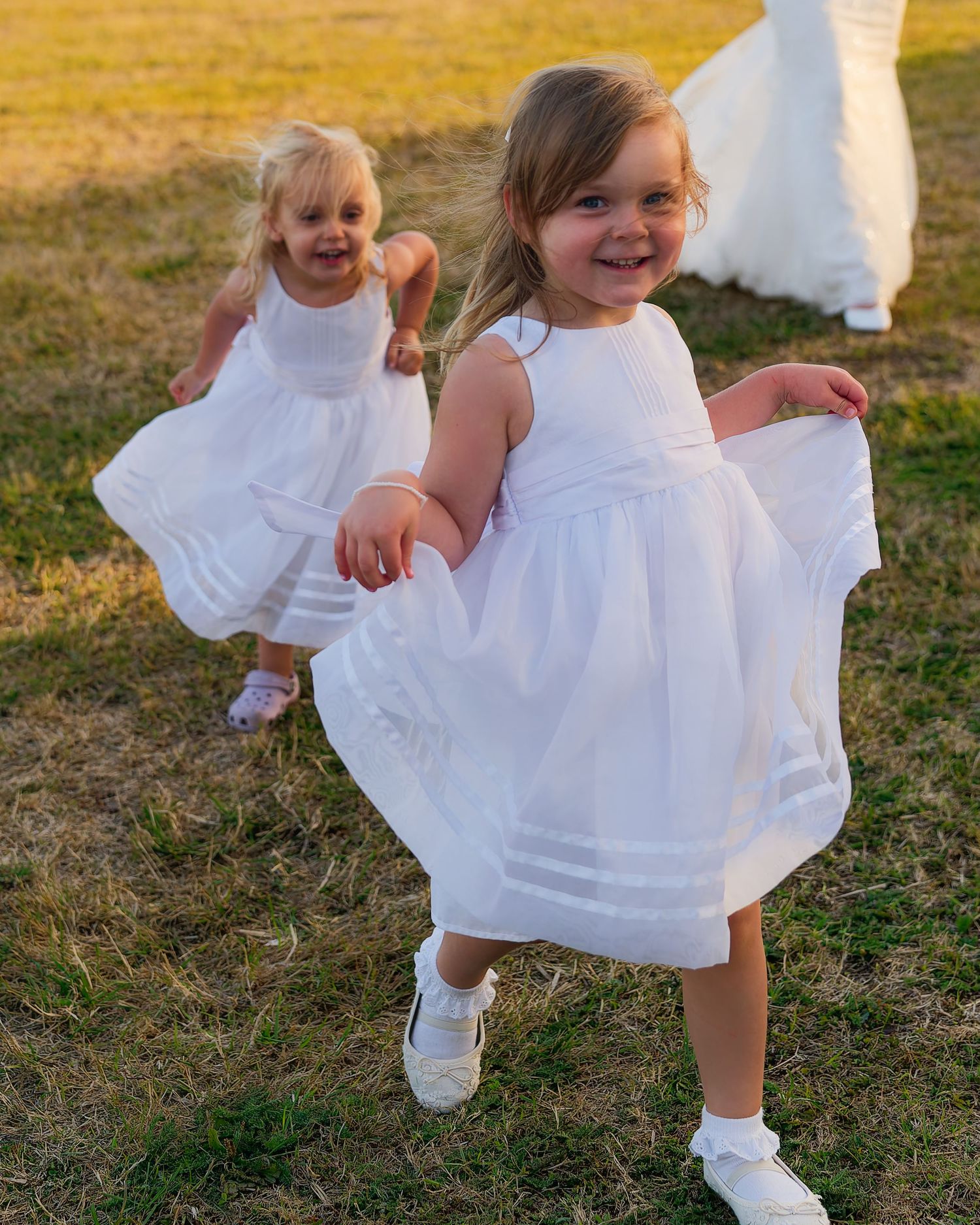 Two young flower girls in white dresses twirling and laughing on a sunlit grass lawn.