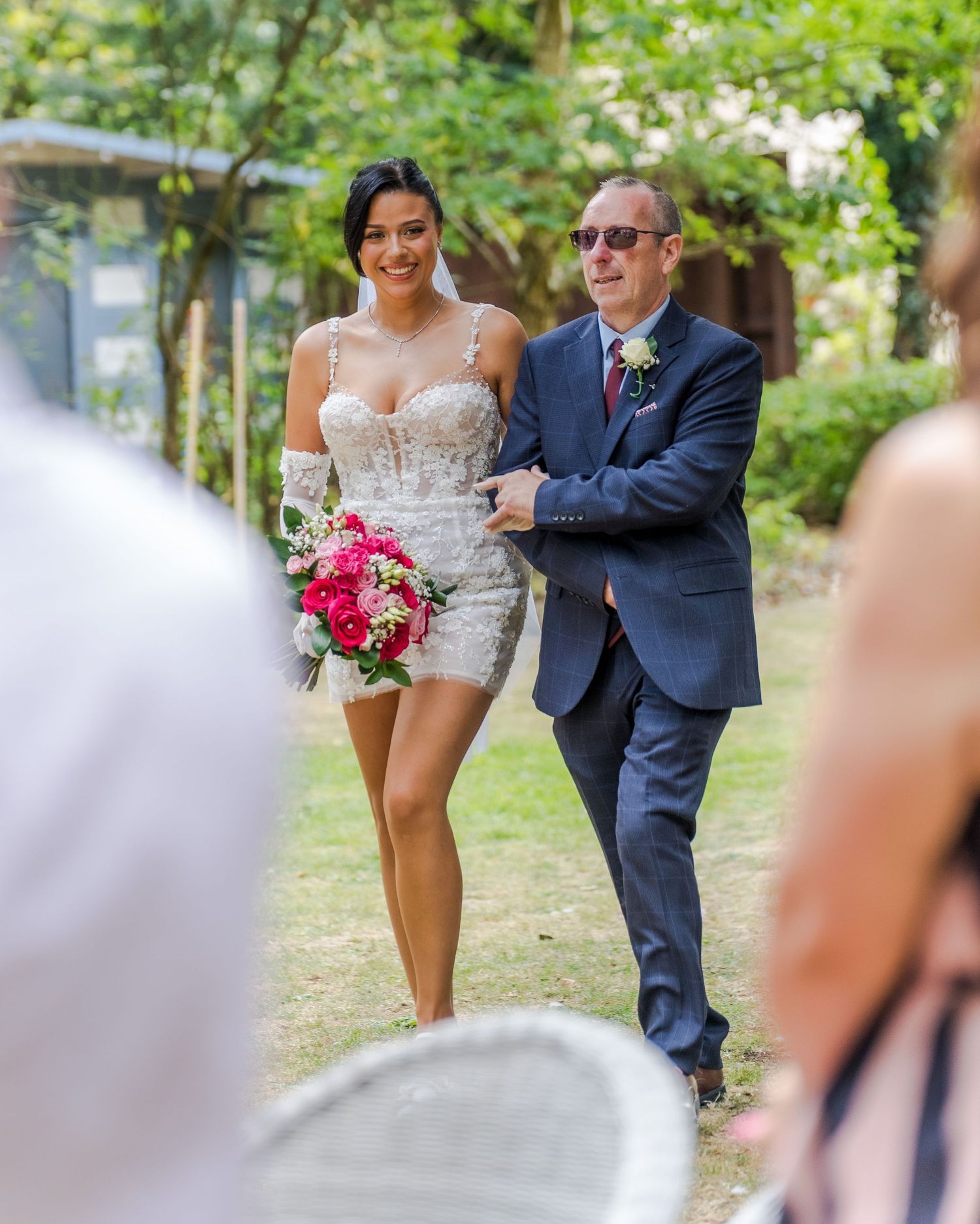 Stunning bride in short lace dress holding pink bouquet walking down aisle with escort.
