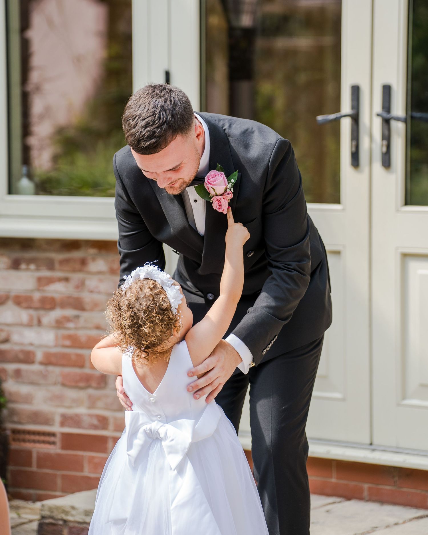 Groom in black tuxedo bending down to greet young flower girl in white dress outside wedding venue.