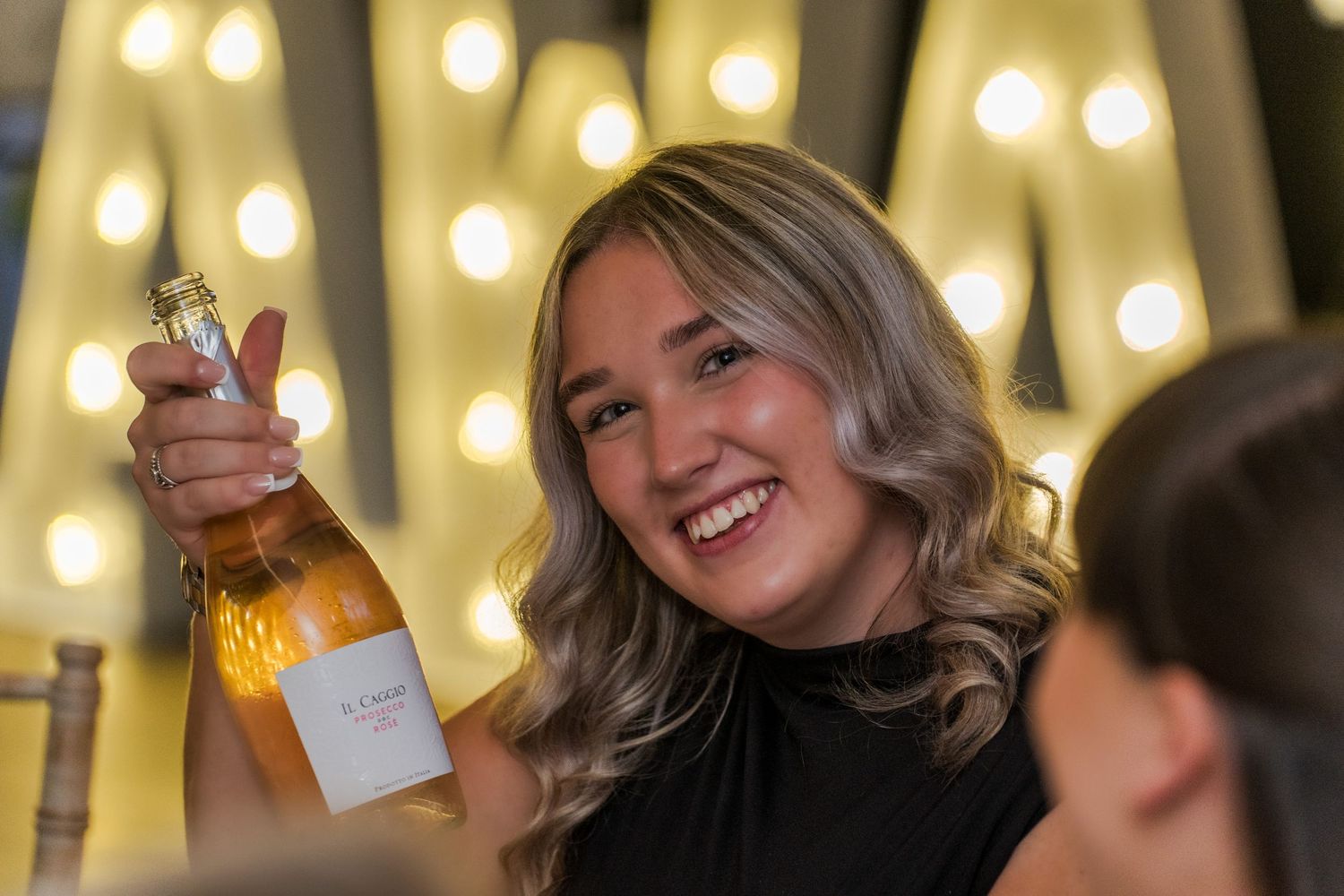 Smiling wedding guest holding up a bottle of rose champagne in front of glowing letter decorations.