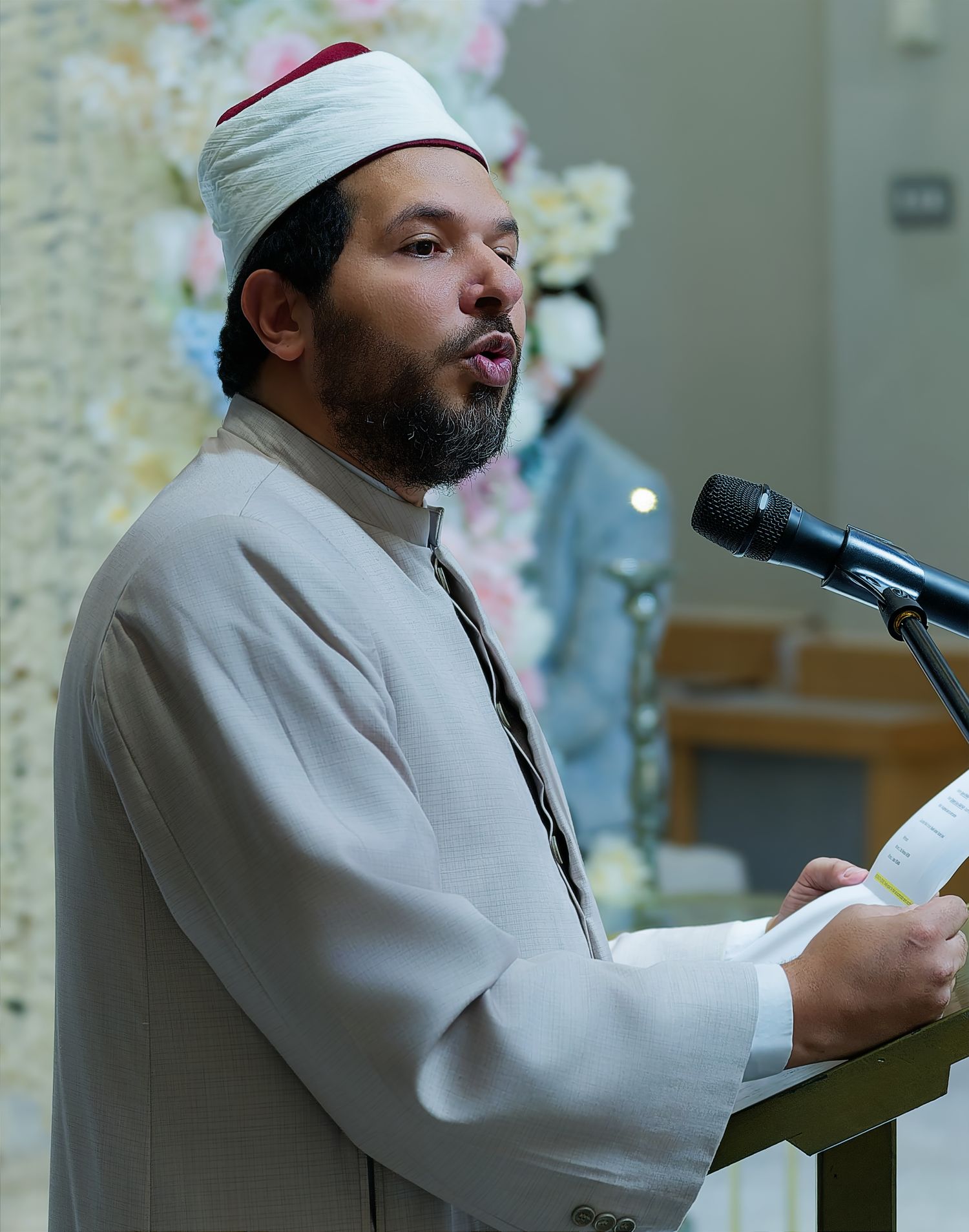 Muslim imam in white robe and red kufi reads from paper at wedding ceremony podium.