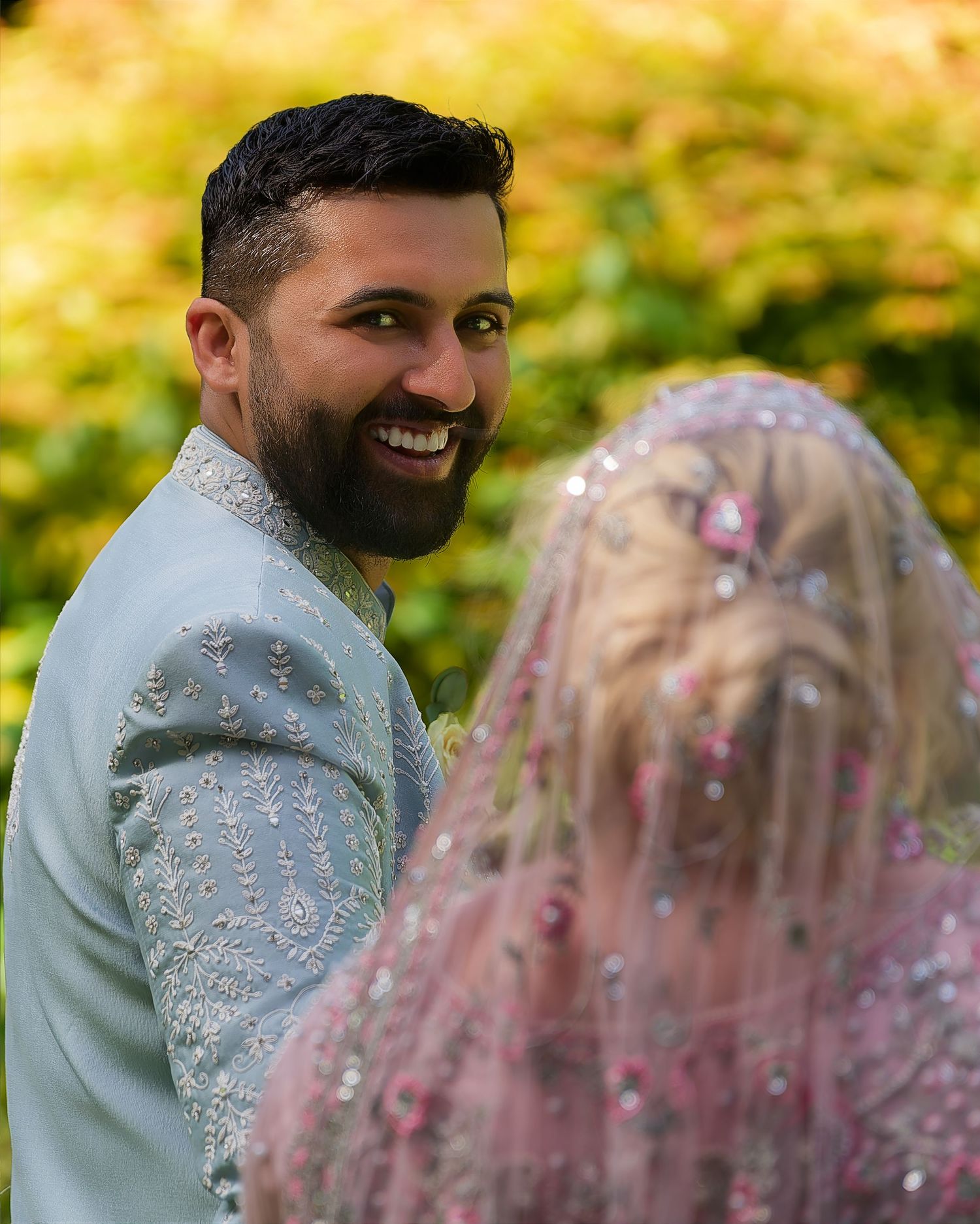A smiling bearded groom in a light blue sherwani looks back at his bride in a pink embellished veil.
