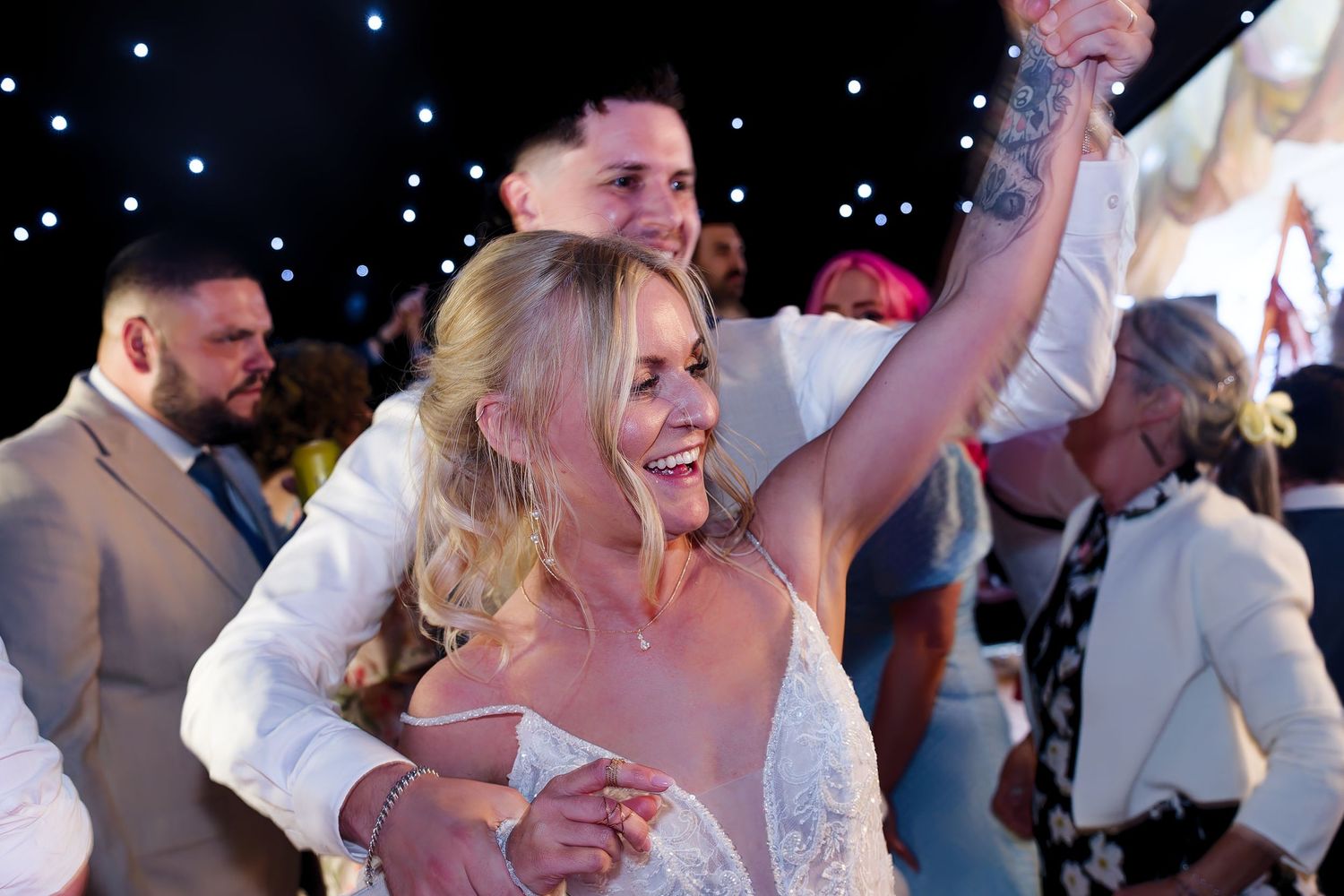 A joyful bride in a white gown raises her arm while dancing with guests at a lively wedding reception.