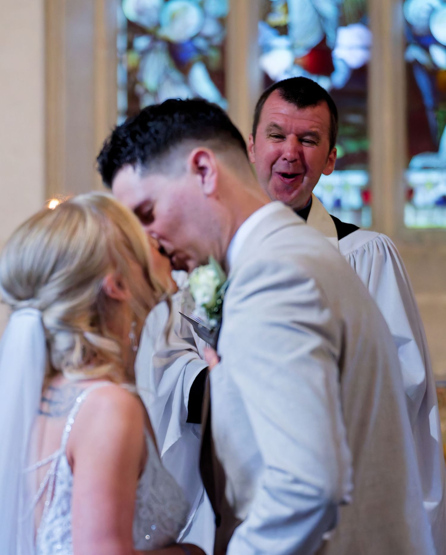 Groom kissing bride at church altar while officiant reacts with joy behind them.