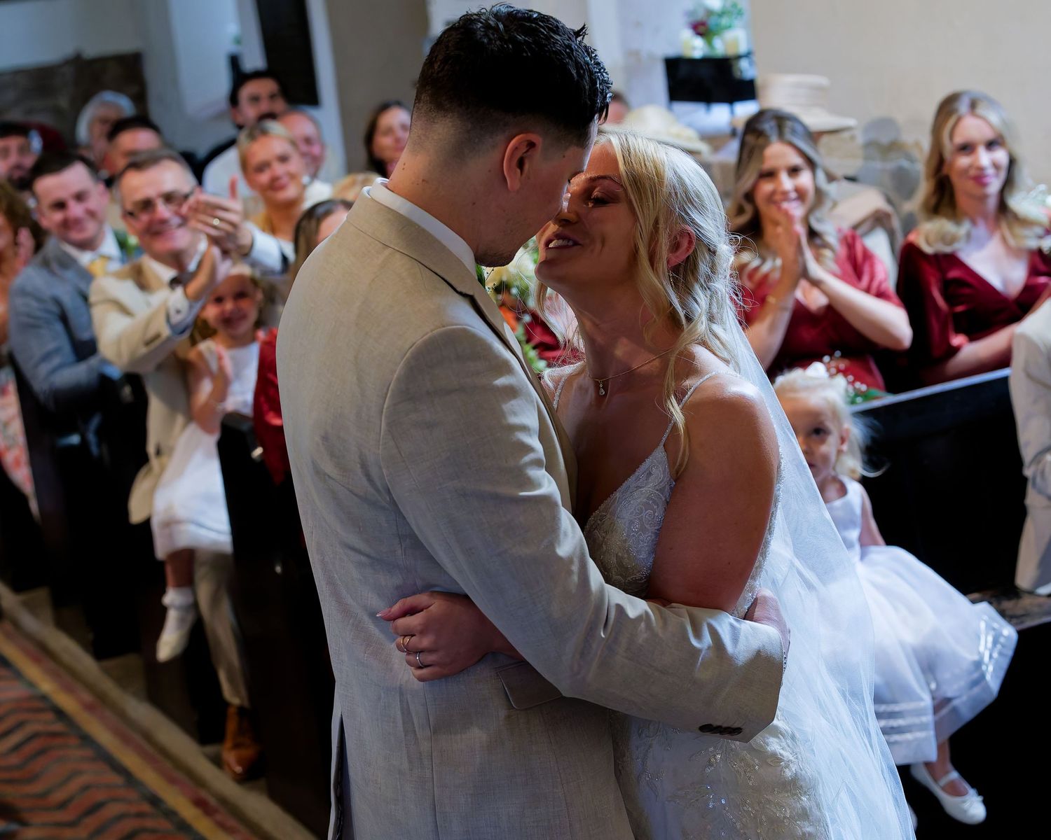 Newlyweds sharing first kiss surrounded by cheering guests at wedding reception.