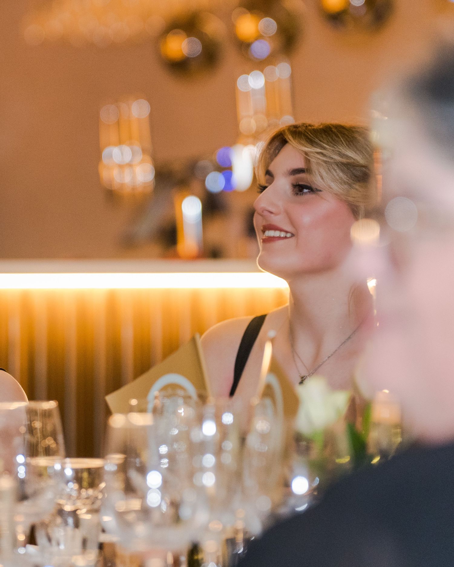 Elegant blonde woman in black dress smiling at wedding reception table with glassware and warm lighting.