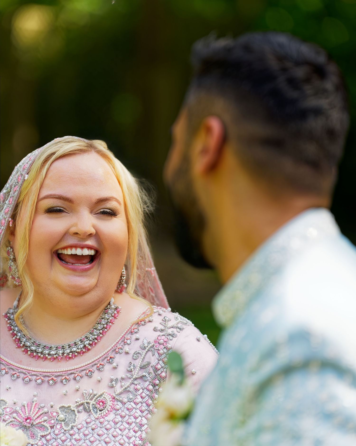 Blonde bride in pink embroidered South Asian bridal outfit laughing joyfully during outdoor wedding ceremony.