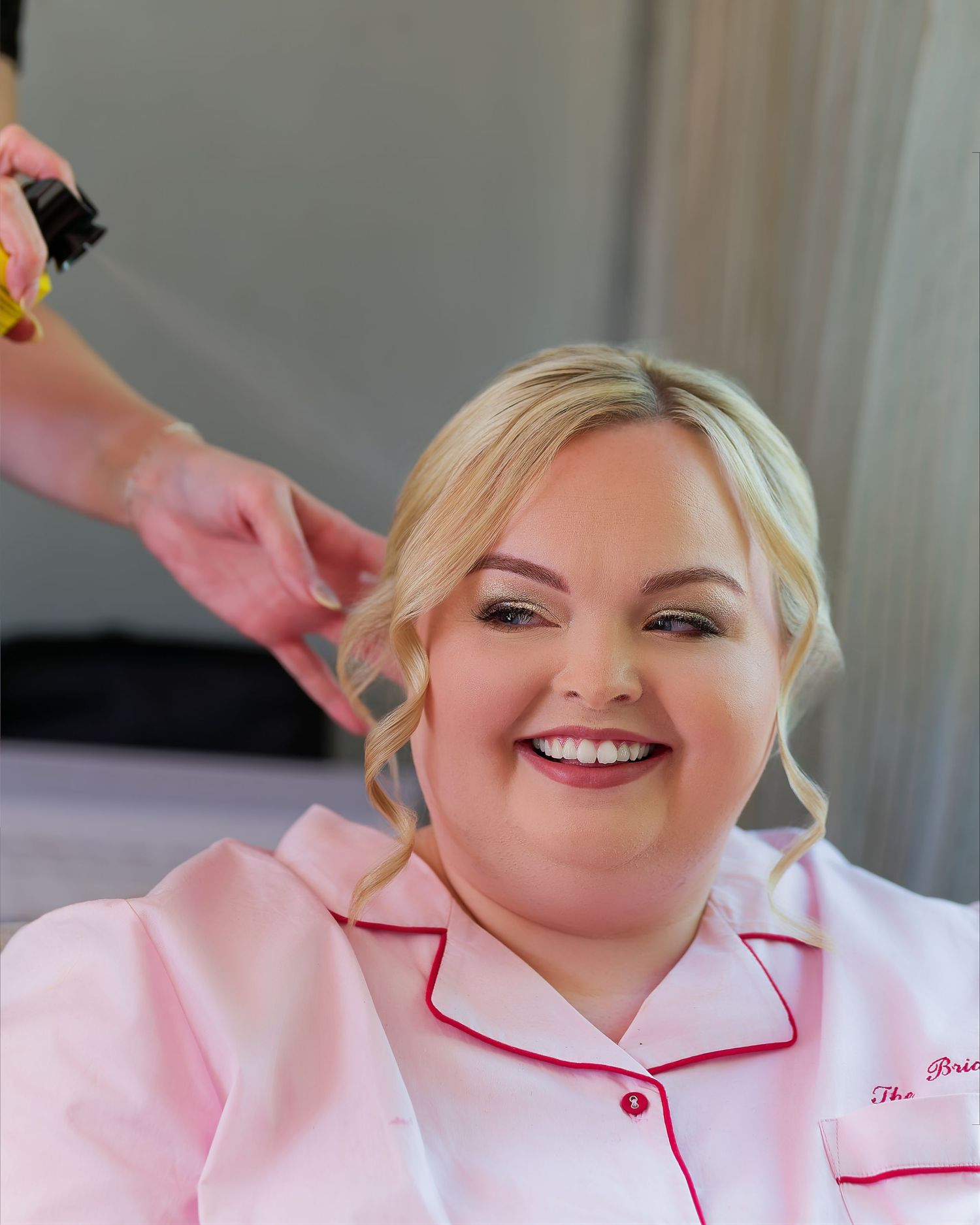 Smiling bride in pink satin pajamas getting her hair styled on her wedding morning.