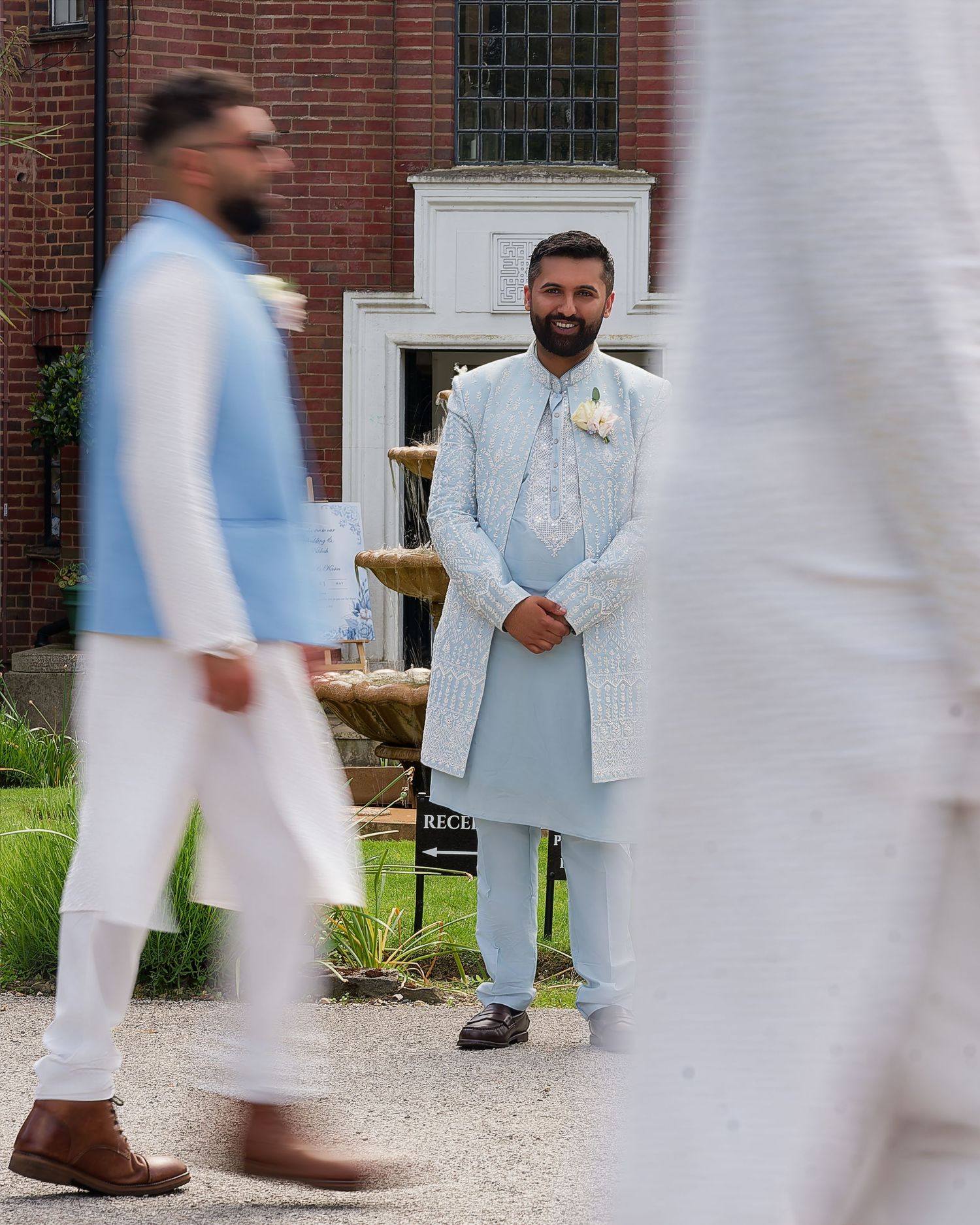Groom in light blue traditional Indian wedding attire waits outside a brick building on wedding day.