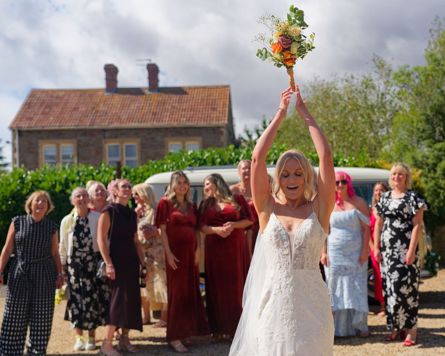 Blonde bride in lace gown tosses bouquet to guests at outdoor wedding with bridesmaids in burgundy.