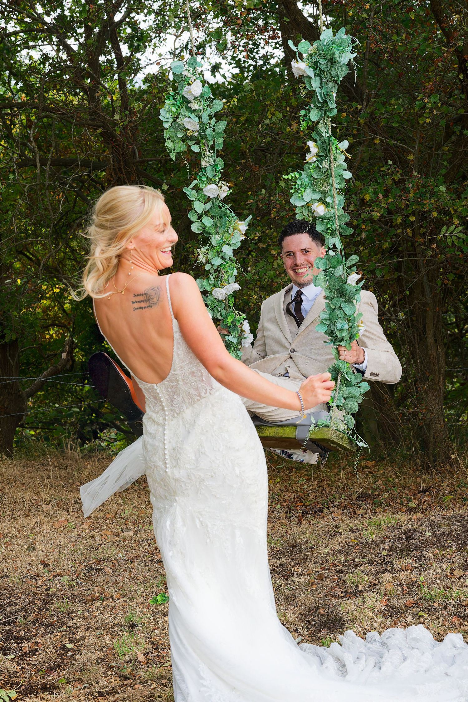 Bride in lace gown pushes groom on floral swing decorated with eucalyptus in woodland setting.