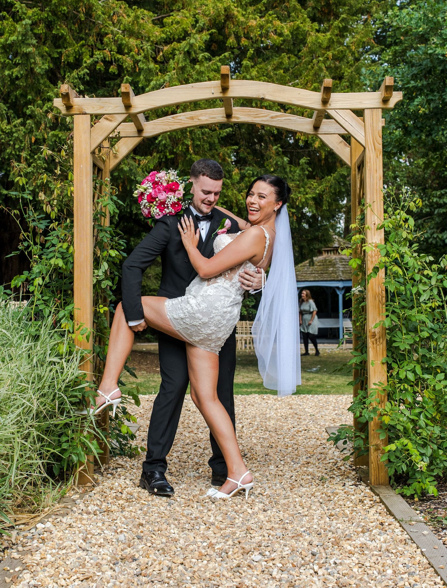Groom lifting laughing bride in short white dress under wooden arch in garden wedding setting.