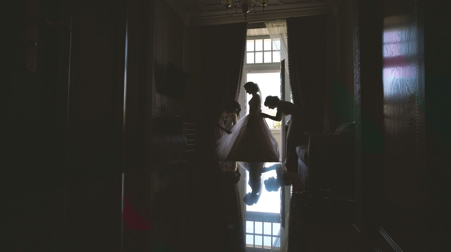 Silhouette of bride in white dress standing in doorway with soft backlight in dark interior.