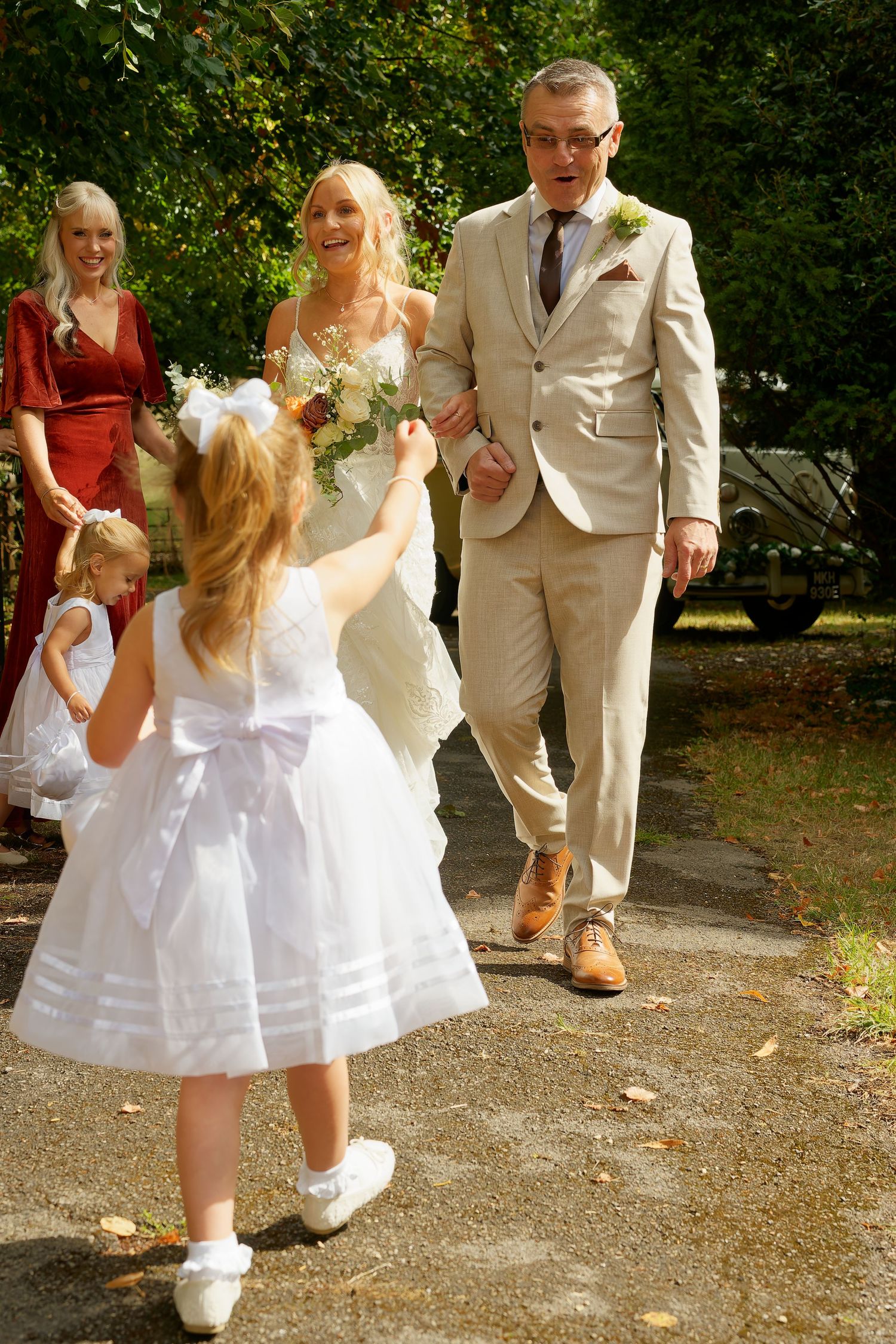 Bride and groom walking outdoor path as flower girl in white dress runs to greet them.