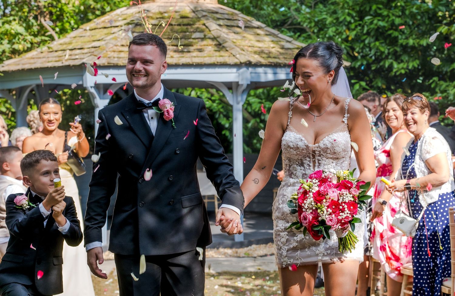 Newlywed couple walks hand in hand as guests throw red confetti near a garden gazebo.