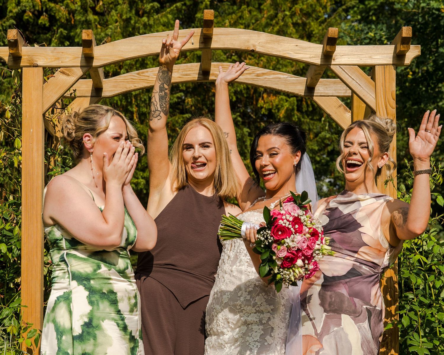 Bride with pink bouquet celebrates joyfully with three bridesmaids under wooden arch in garden.