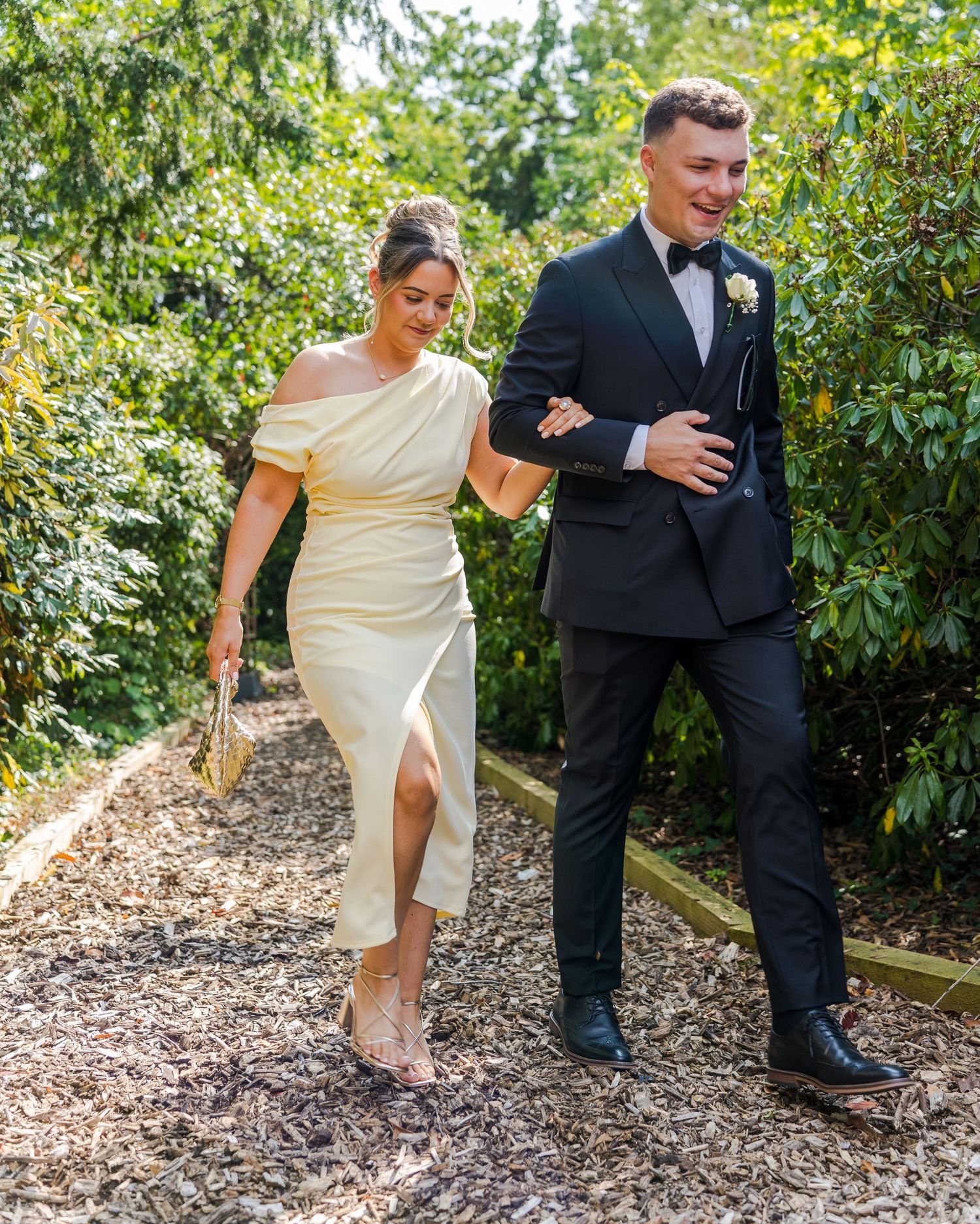 Elegant wedding guests walking garden path, woman in ivory off-shoulder dress, man in tuxedo.