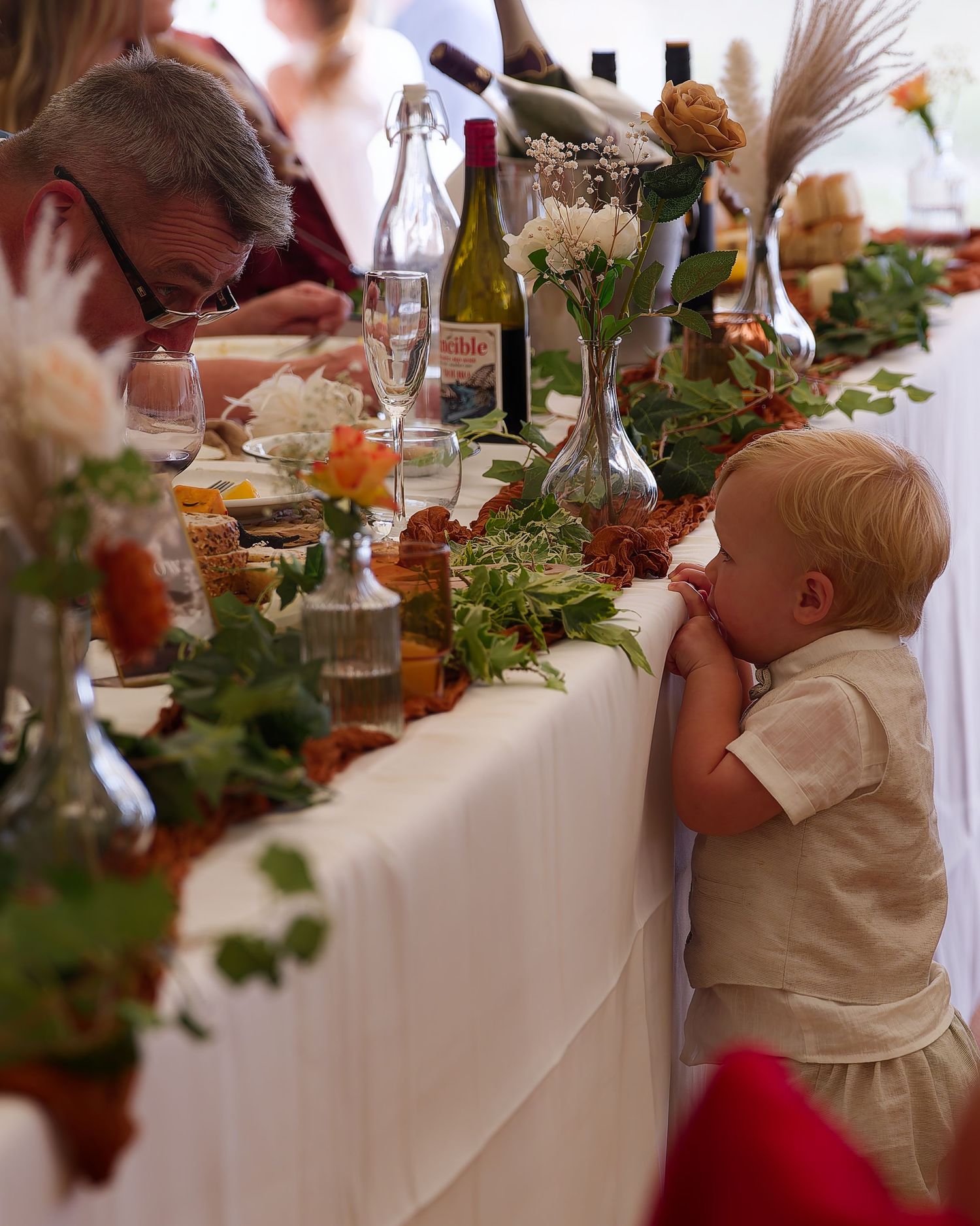 Adorable toddler in a linen outfit reaching up to peek at a beautifully decorated wedding table.