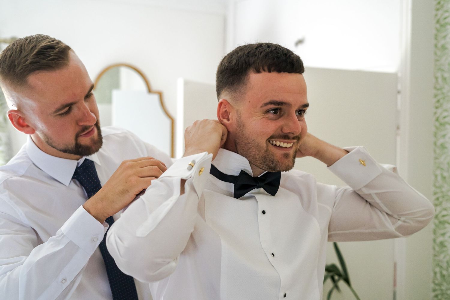 Groomsman helping groom adjust black bow tie while smiling during wedding preparations.
