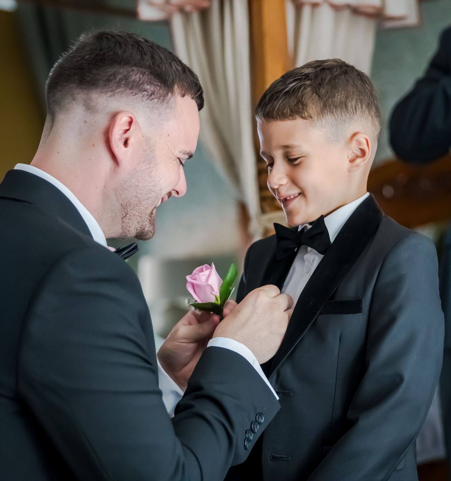 Groom in gray suit pins a pink rose boutonniere on a young boy in a tuxedo.