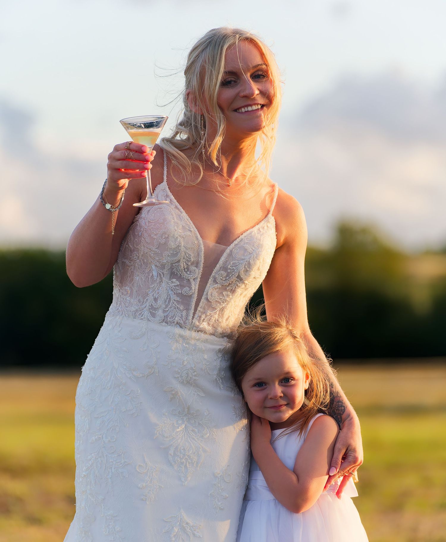 Smiling blonde bride in lace gown holds cocktail glass while young flower girl hugs her at sunset.